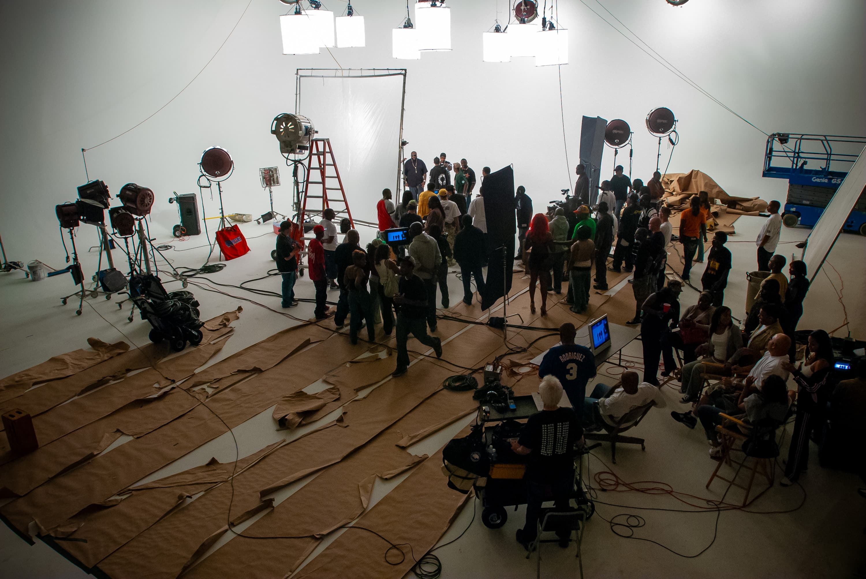 A wide angle view of the set of the music video "Down In Tha Dirty". The cast and crew is working on a white cyclorama wall with tattered craft paper taped to the floor. There are various lights and tools for filmmaking scatter around the studio.