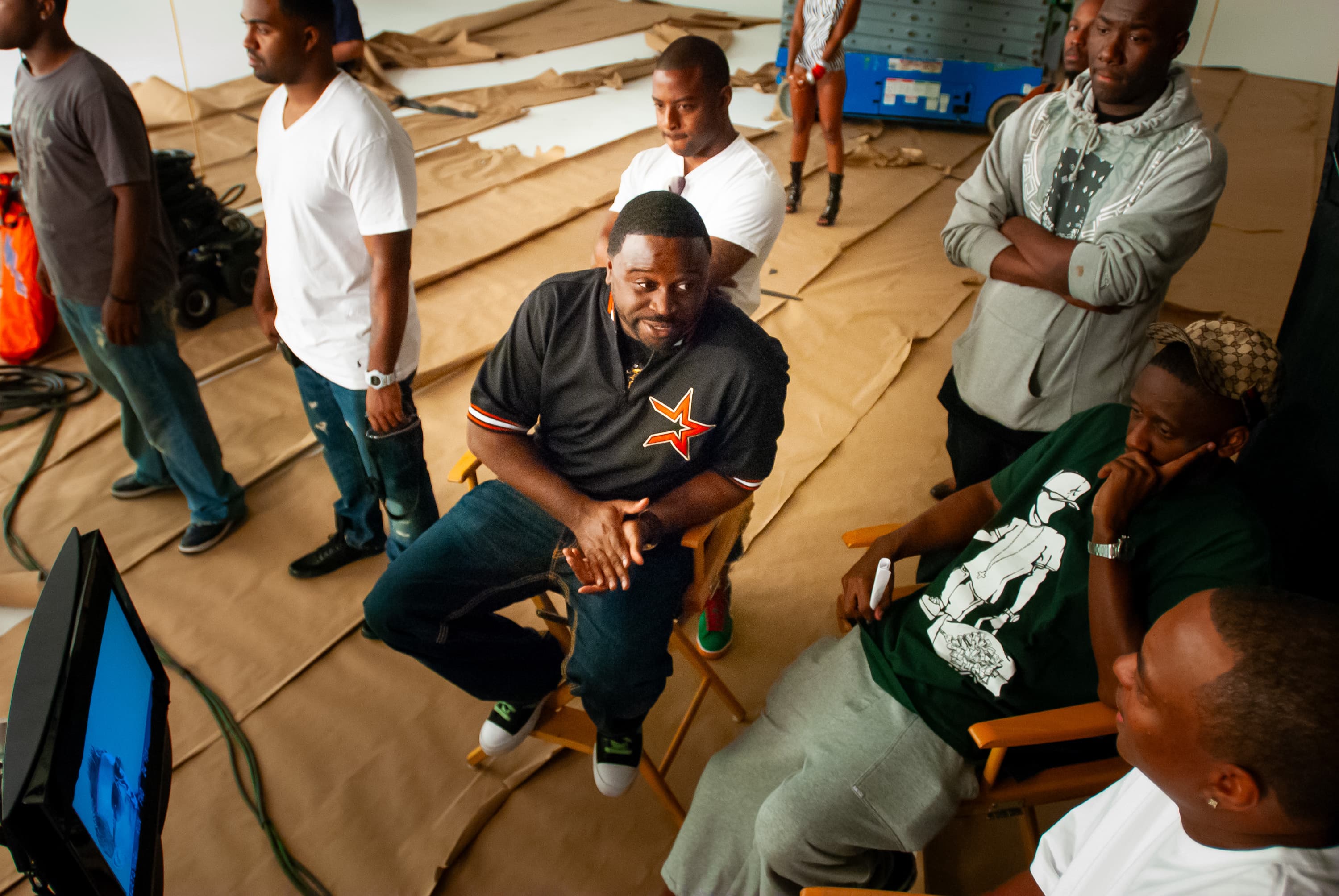 Director Dr. Teeth sitting in a directors chair on the set of a music video, circled around a video monitor with other crew and talent. The floor is covered with torn craft paper.