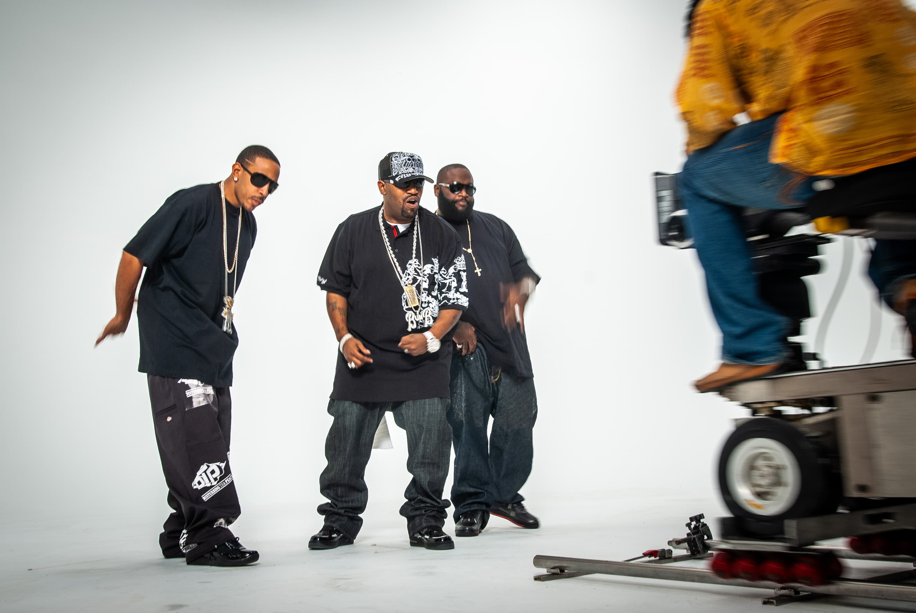 Rappers Ludacris, Bun B and Rick Ross performing on a white featureless background while a movie camera on a dolly rolls towards them.