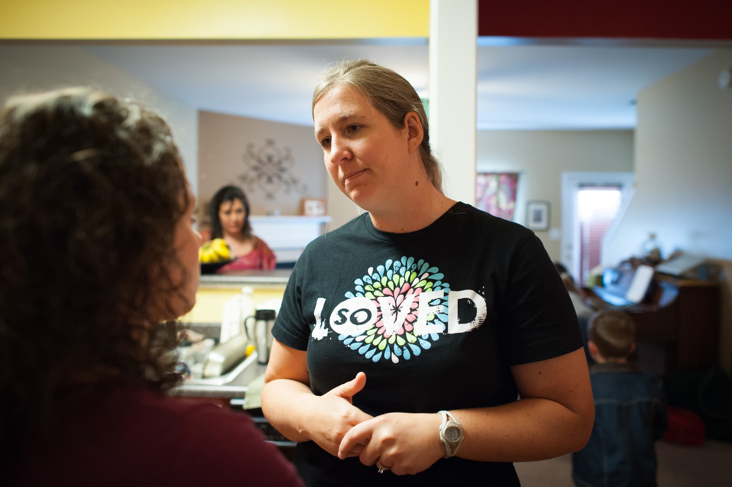 A woman wearing a shirt that says "so LOVED" stands in a room, talking to another person. In the background, a woman is in the kitchen and a child is near a piano.