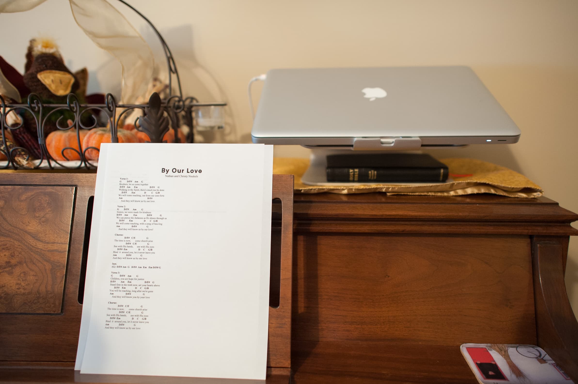 A sheet of paper with song lyrics titled "By Our Love" by Nathan and Chrissy Nockels on a wooden surface. A laptop and a book are on the right side, resting on the same surface.