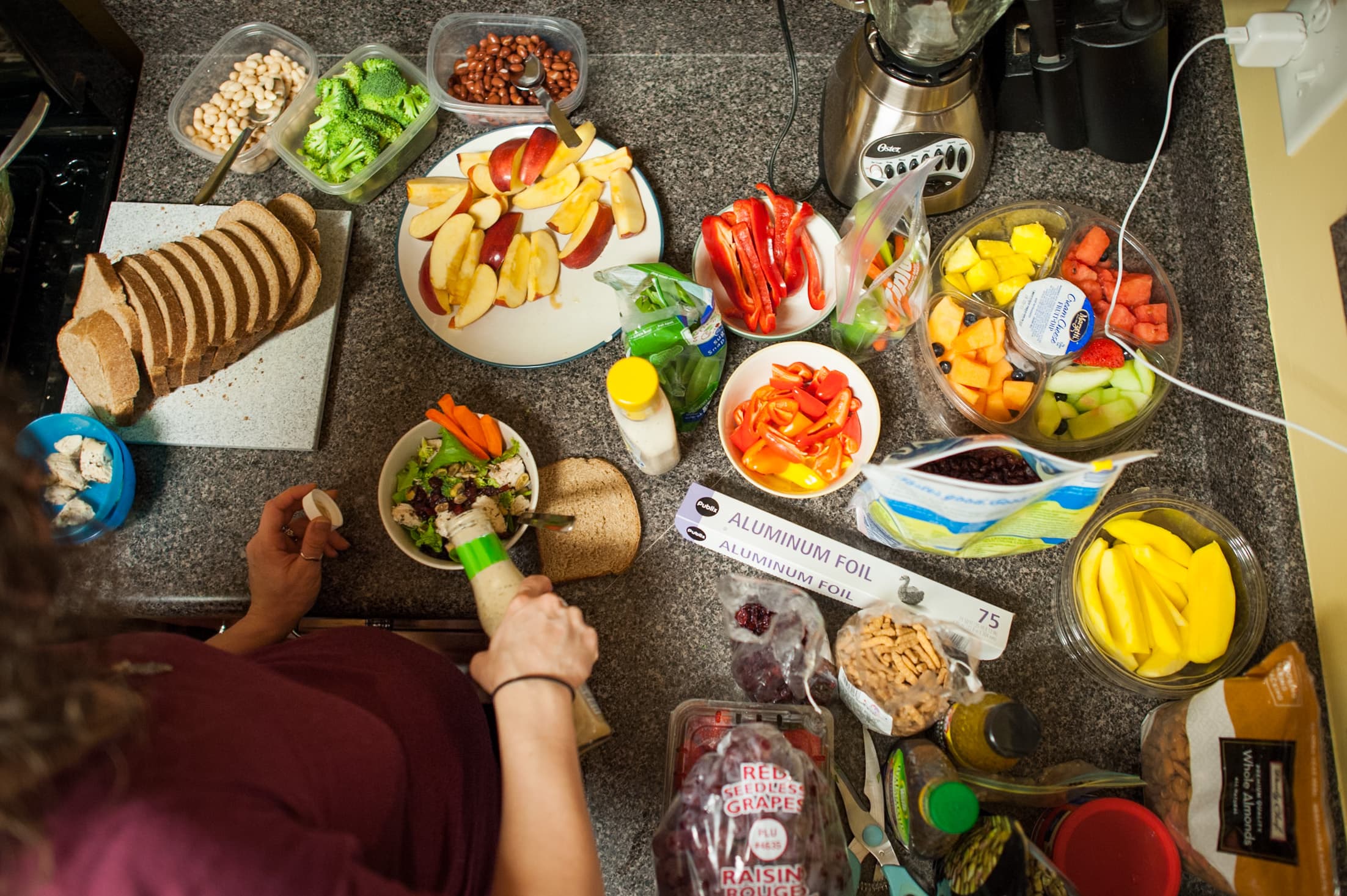 A kitchen countertop with various food items, including sliced bread, broccoli, beans, apple slices, red bell peppers, carrot sticks, and a fruit mix container. A person is preparing a meal with salad and dressing. Other items include aluminum foil, red grapes, and a blender.