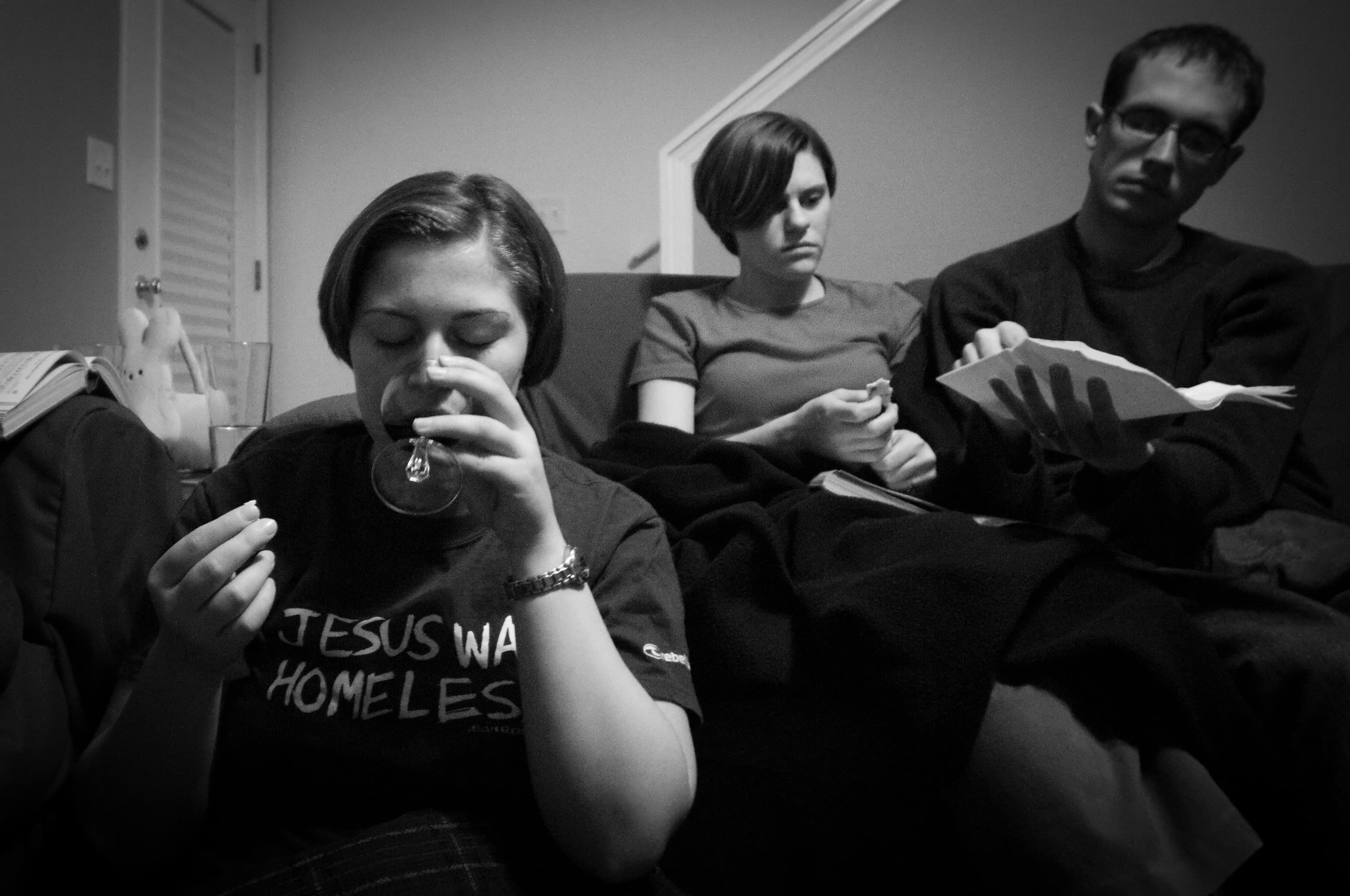 A person drinks from a glass with another person holding food next to them, and a third person holding an open book. The front person's shirt reads "JESUS WAS HOMELESS."