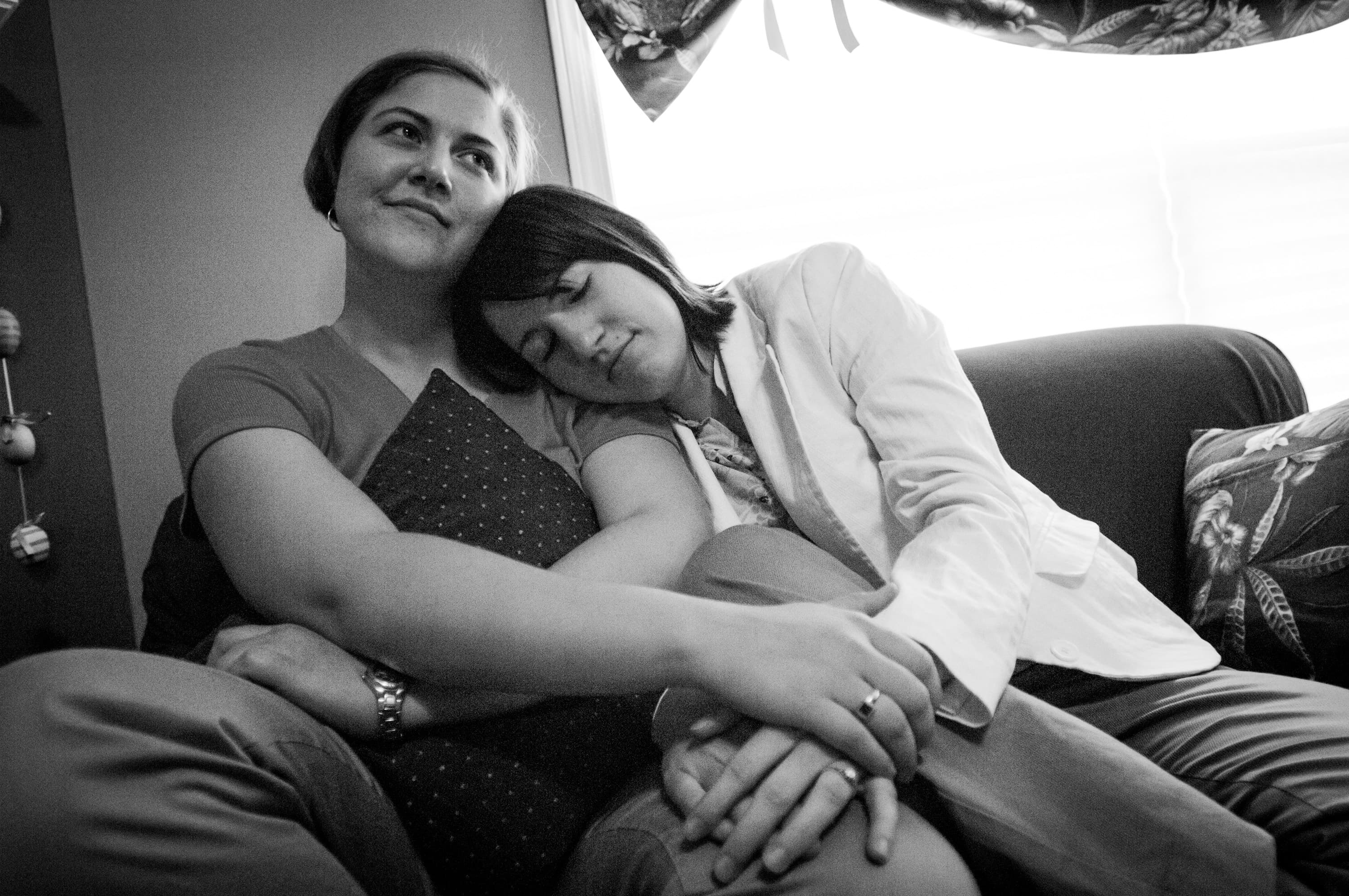 Two people sitting on a couch. One person is leaning on the other's shoulder with their eyes closed, while the other person is looking upward with a slight smile, holding a pillow.