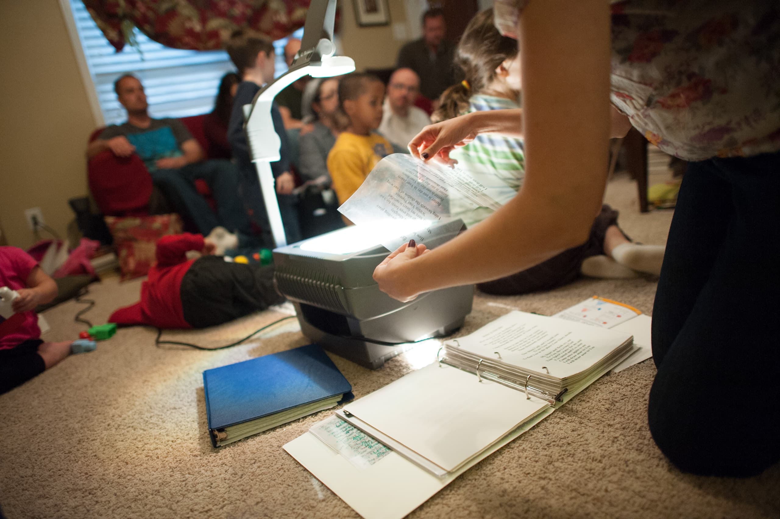 A person using an overhead projector with a sheet of text on it. A binder with papers is open on the floor. In the background, several people and children are seated on a couch and the floor.