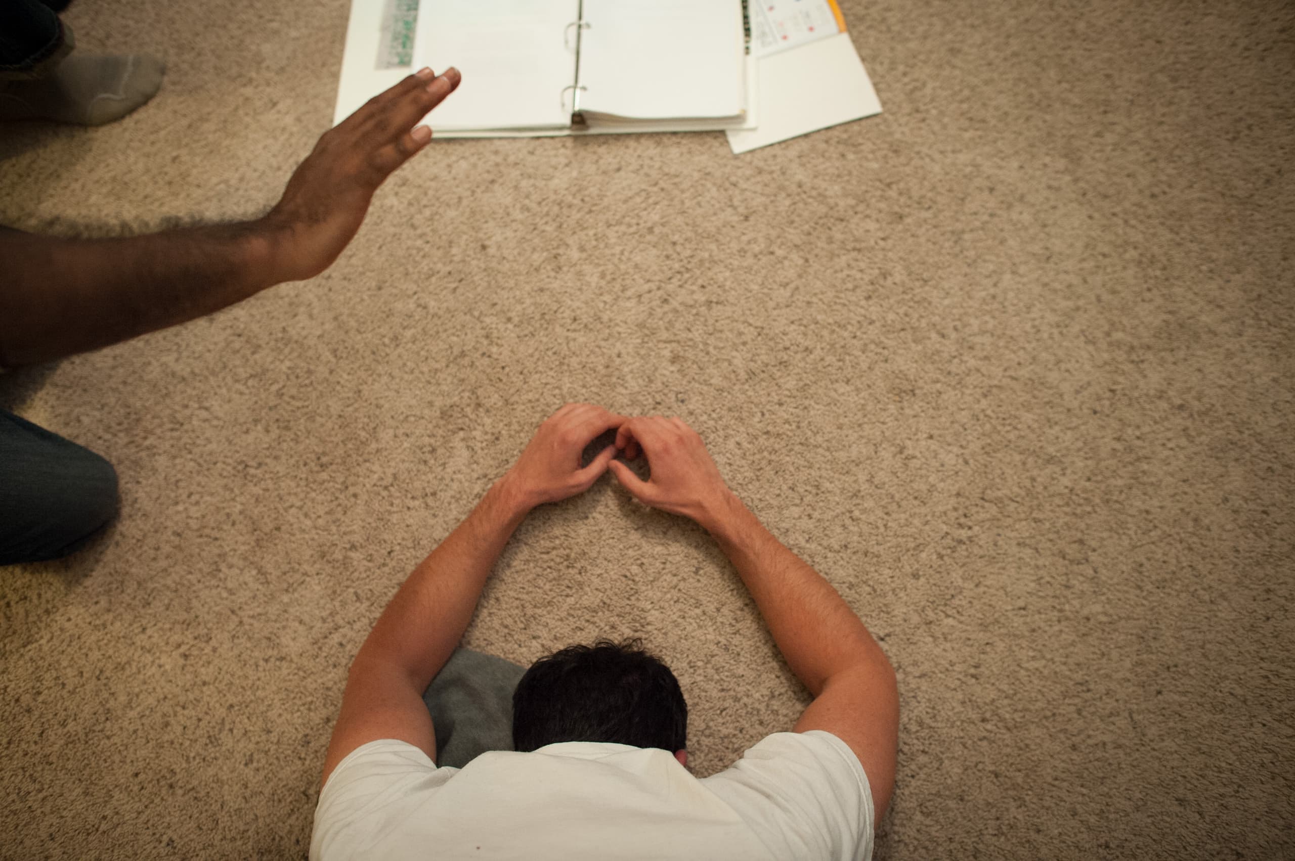 A person lying face down on the carpet with hands on the floor. An arm is extended above them, and an open binder is on the floor nearby.