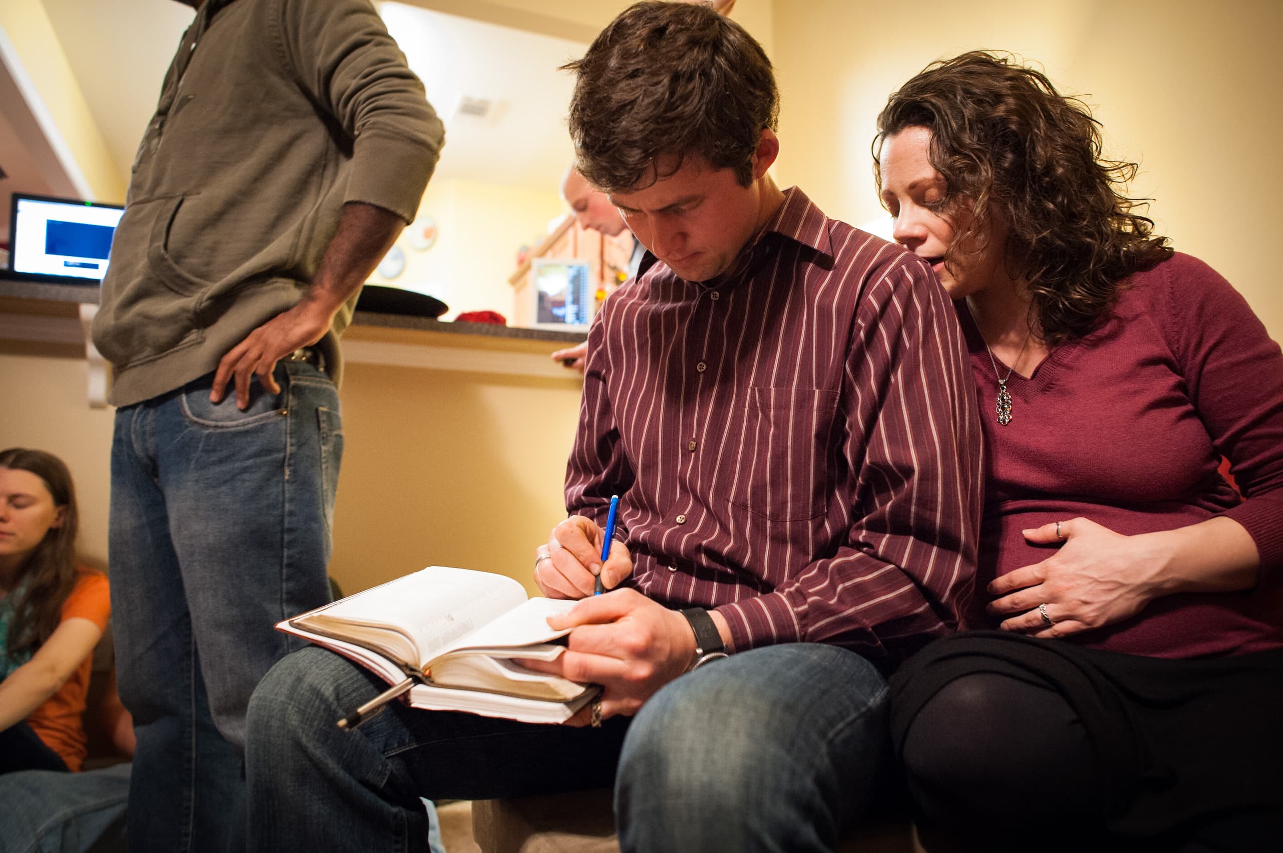 A man writing in a notebook while a woman sits beside him with her hand on her pregnant belly. Other people are visible in the background, including a person standing and another sitting with eyes closed.