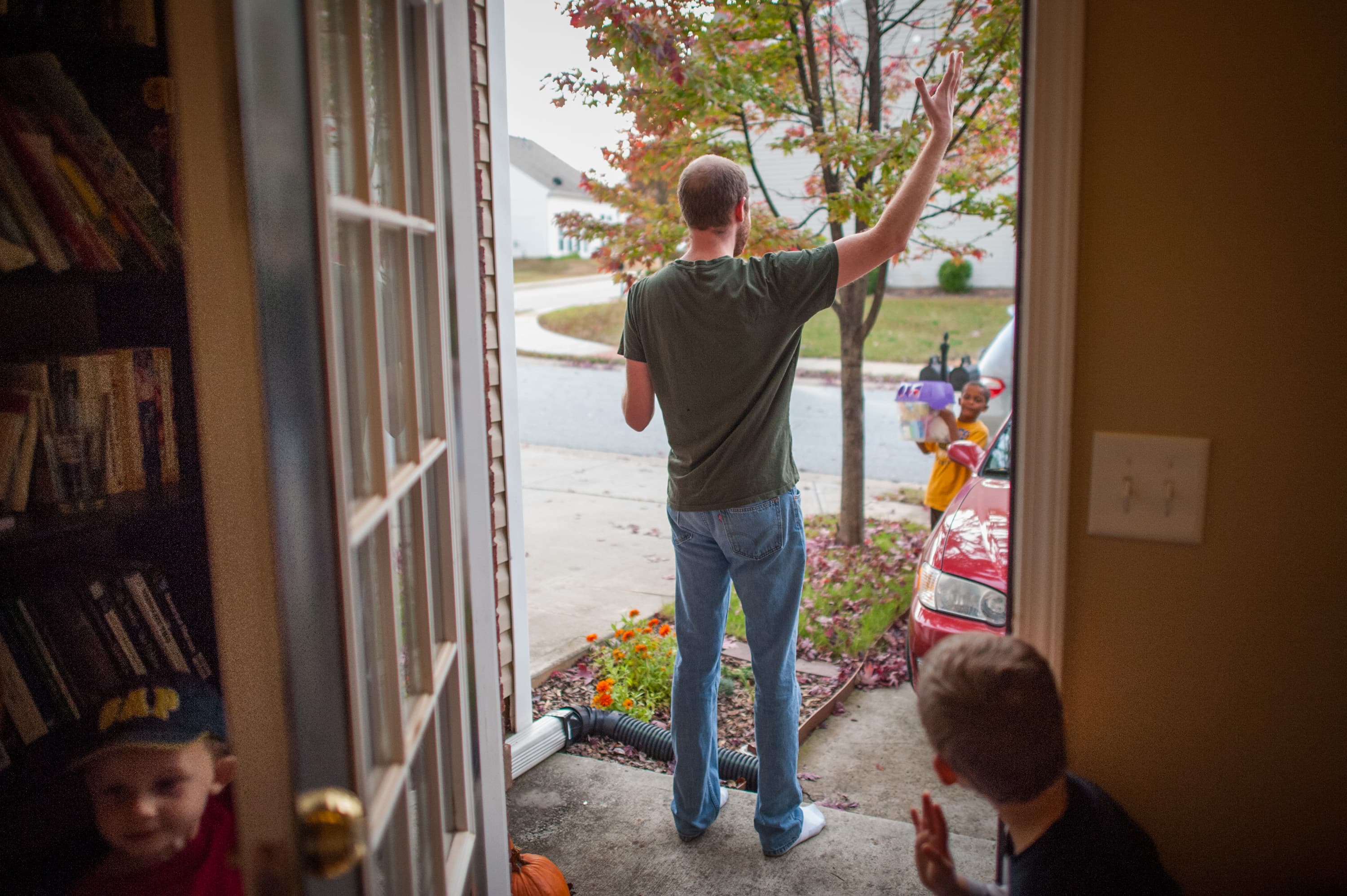 A man stands in a doorway, waving to two children outside near a red car. One child is holding a plastic container. Inside, a shelf of books is visible, and a child in a cap peeks into the doorway.