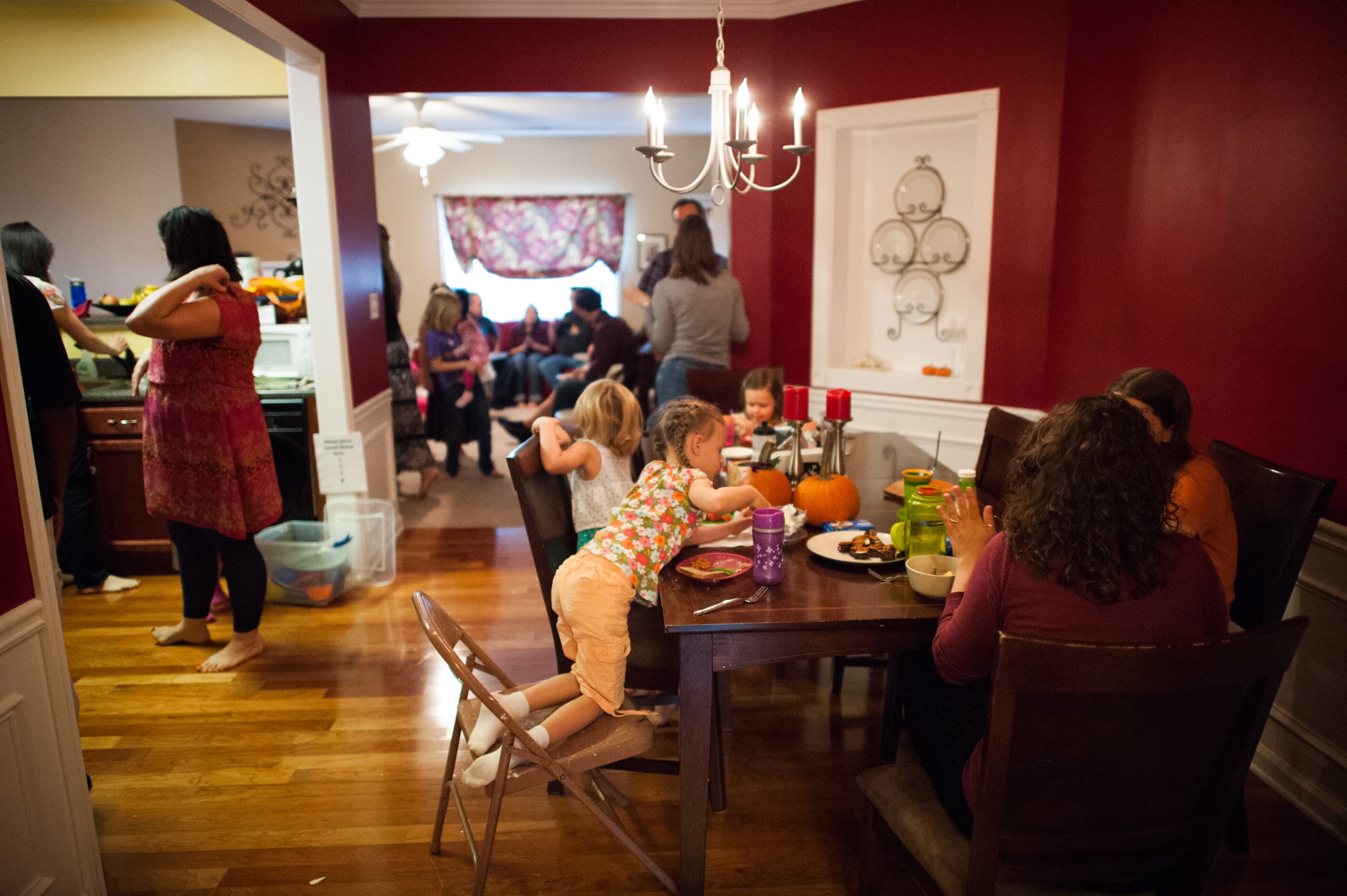 A group of people gathered in a home setting. In the foreground, several children and adults sit around a dining table with food and pumpkins. In the background, more people are gathered in the kitchen and living room area, some standing and others sitting.