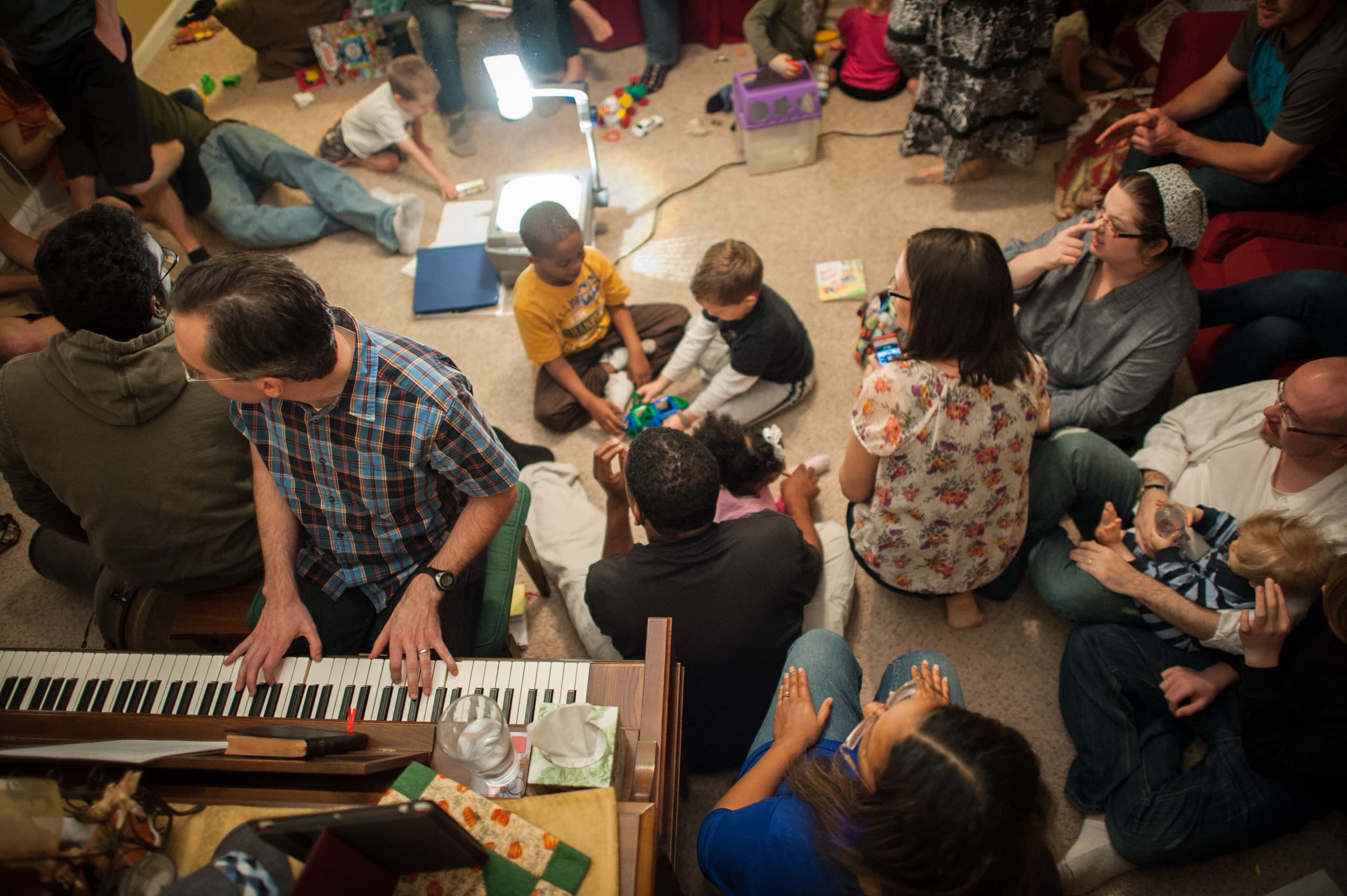 A group of people, including children, gathered in a room. Some are sitting on the floor, and a person is playing a keyboard. Toys and an overhead projector are visible on the floor.