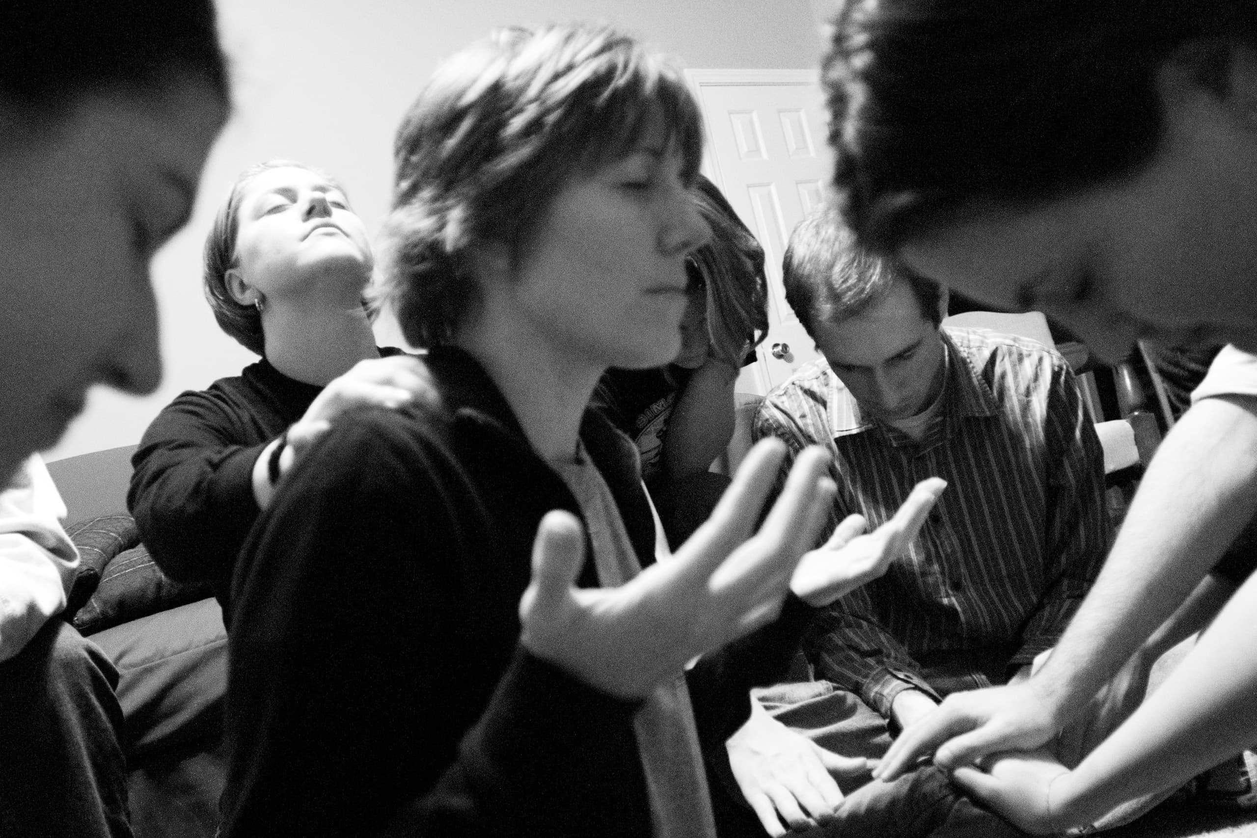 Black and white photo capturing a group of individuals in a moment of collective prayer or reflection. One person is in the foreground with hands open in a gesture of surrender, while others place supportive hands on shoulders, bowing their heads in deep focus.