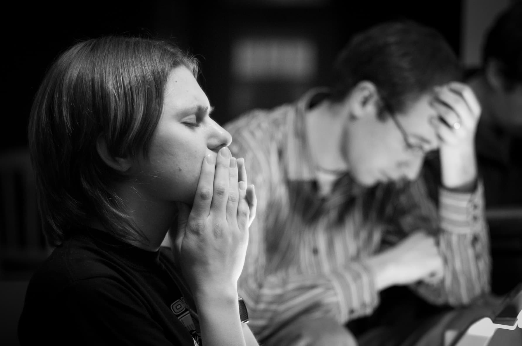 Black and white photo of a person with their hands clasped near their face, appearing deep in thought or prayer, while another person in the background rests their head on their hand, also reflecting a contemplative mood.