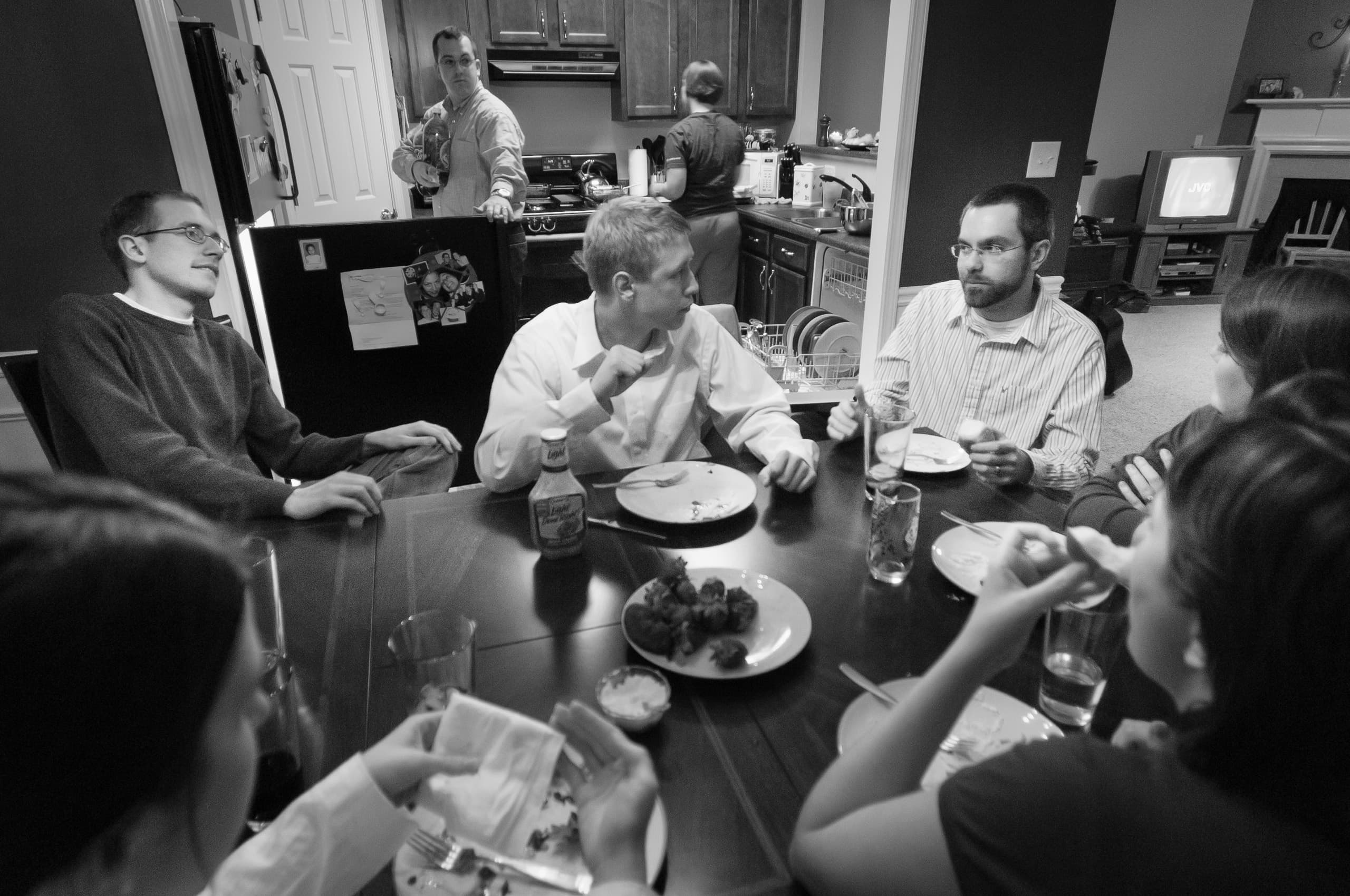 Black and white photo of a group of people gathered around a dining table, sharing a meal and engaged in conversation. In the background, a couple of individuals are in the kitchen, adding to the lively, communal atmosphere.