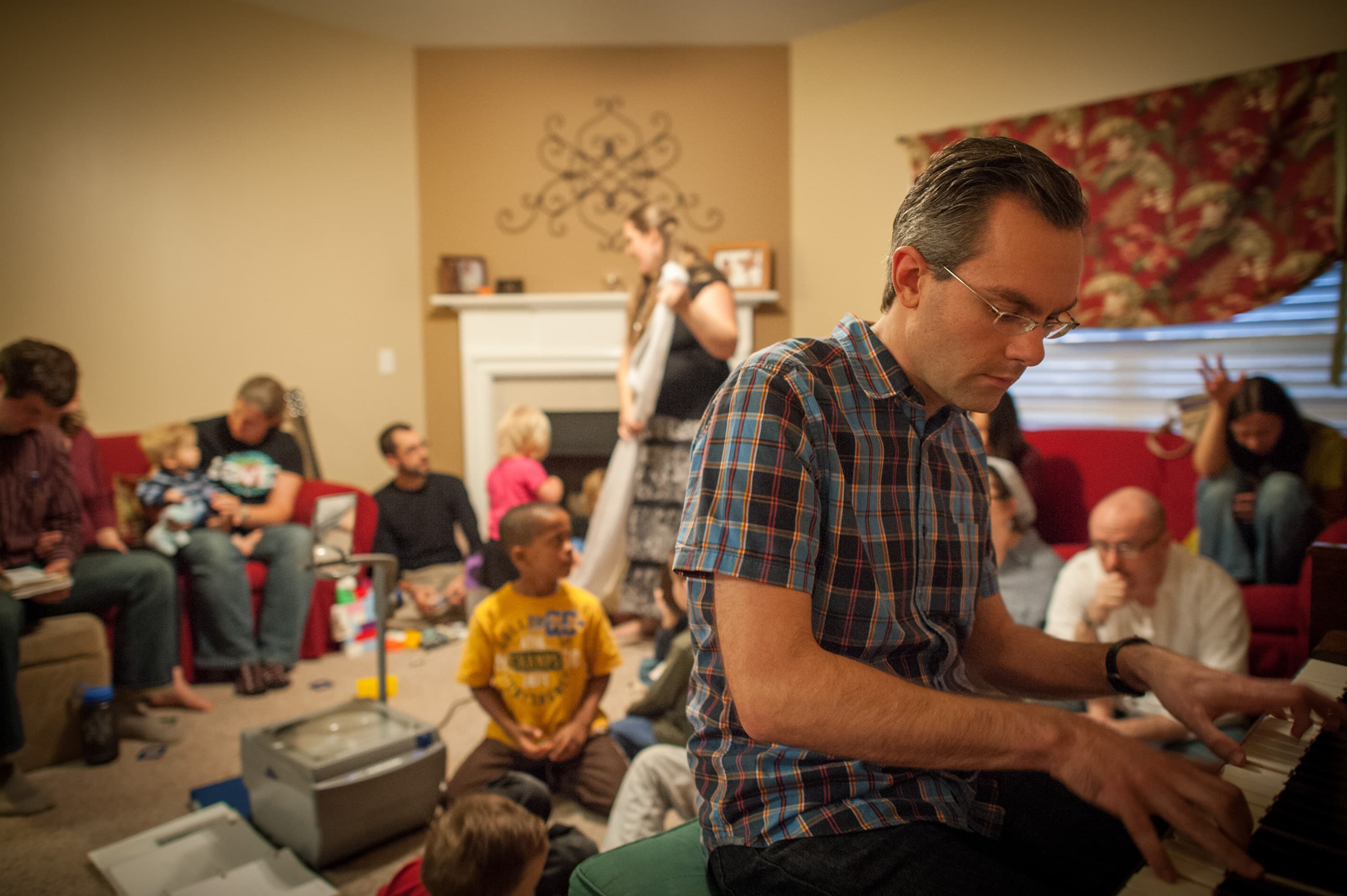 A group of people gathered in a living room. A man in the foreground is playing a piano. Other adults and children are sitting or standing around the room, engaging in various activities. Some are seated on couches, while others are on the floor. A fireplace and decorated wall are visible in the background.