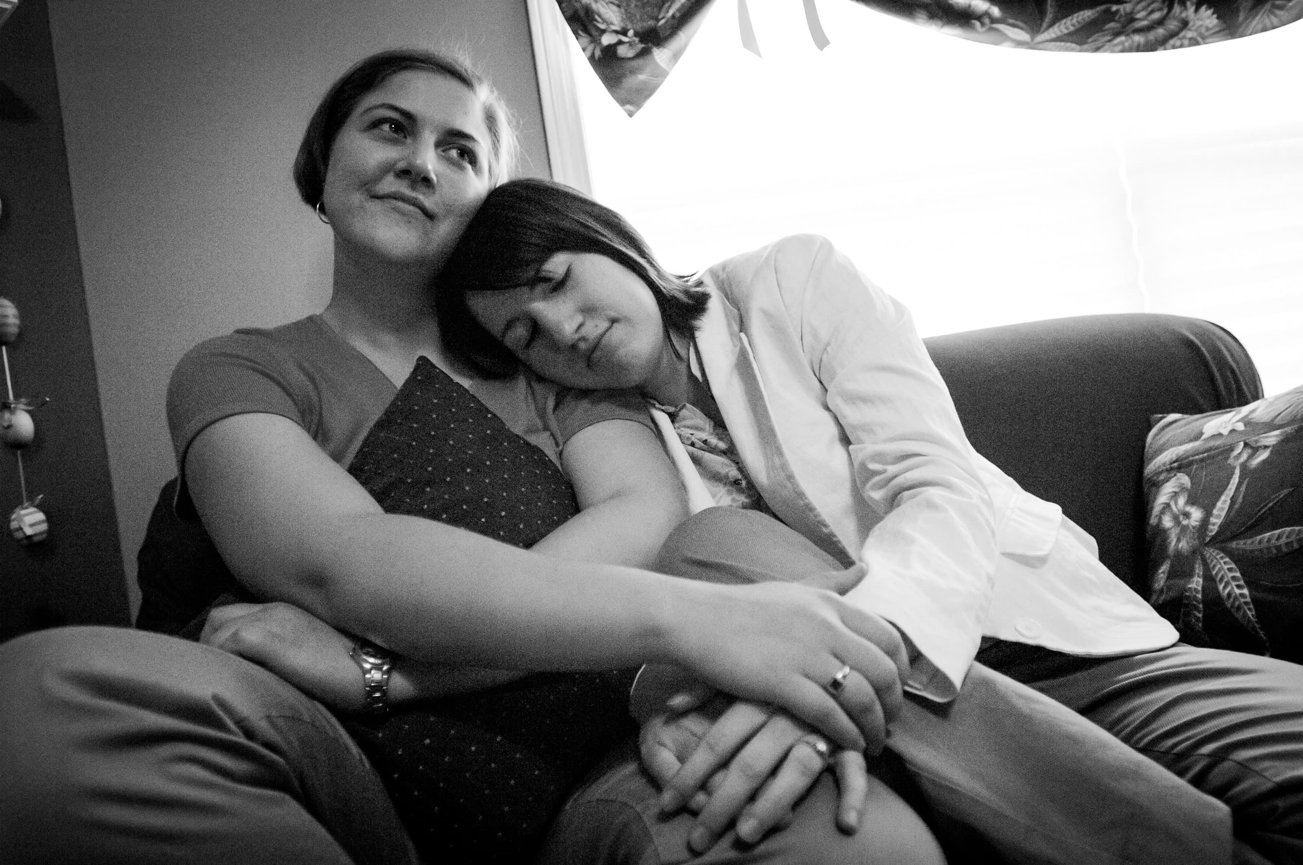 Two people sitting on a couch. One person is leaning on the other's shoulder with their eyes closed, while the other person is looking upward with a slight smile, holding a pillow.