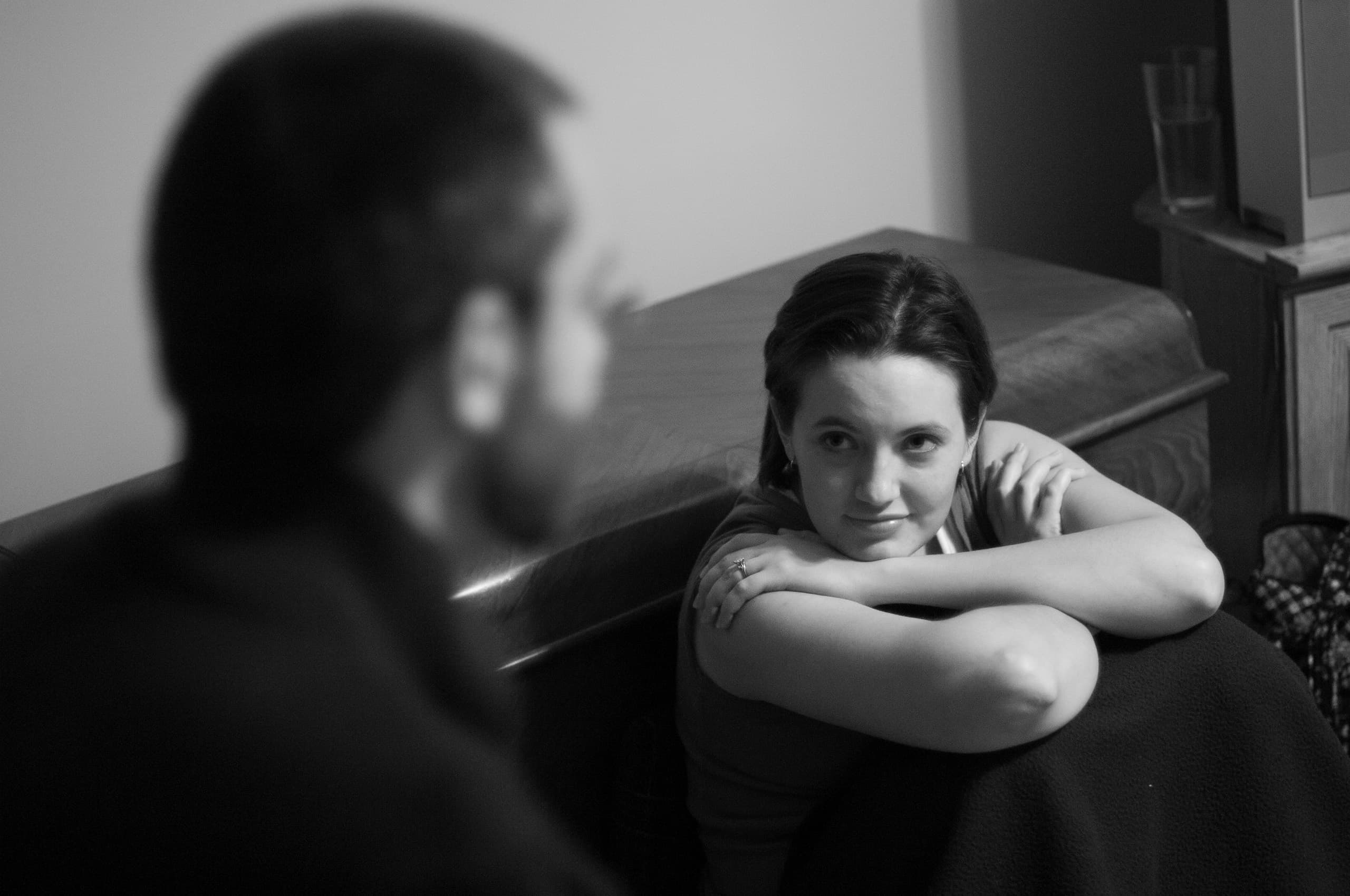 A woman leaning on her arms next to a piano, facing a man who is out of focus. A glass is on a table in the background.