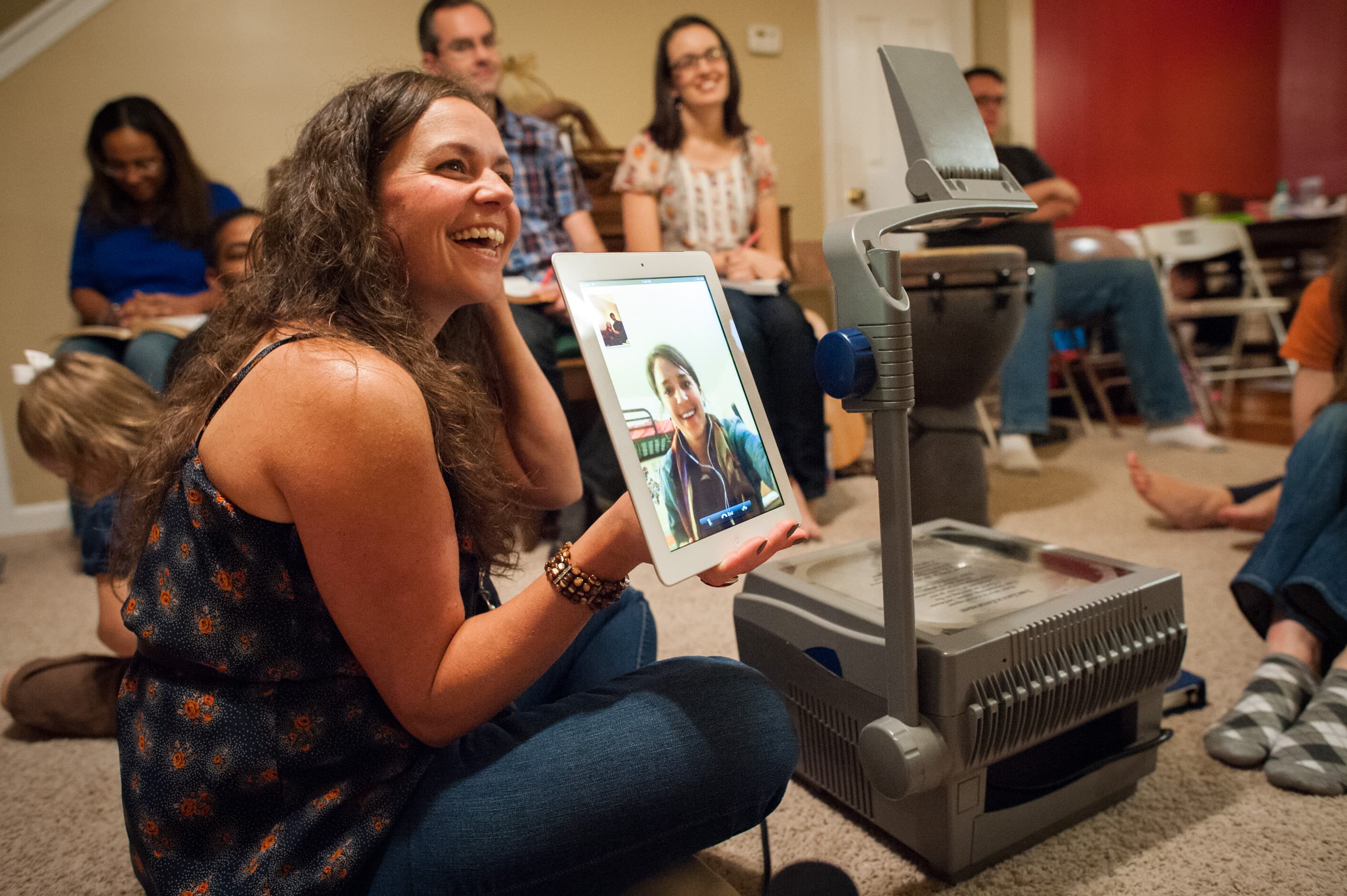 A woman holding a tablet with a video call on the screen, sitting next to an overhead projector in a room with several people gathered around. Some people are seated on chairs, while others are sitting on the floor.