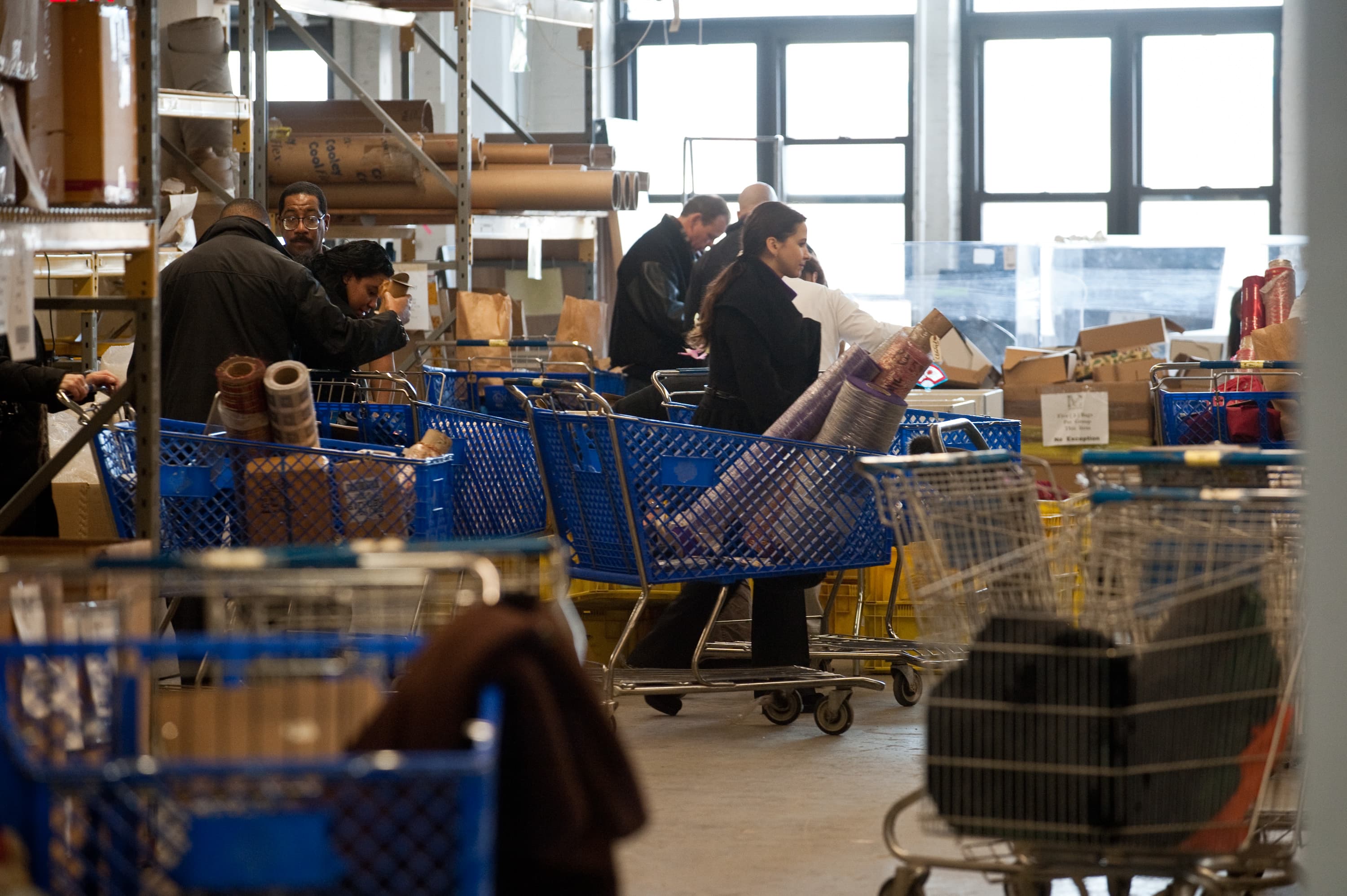 People with shopping carts in a warehouse-like setting, looking at various items, including rolls of material. Shelving and windows are visible in the background.