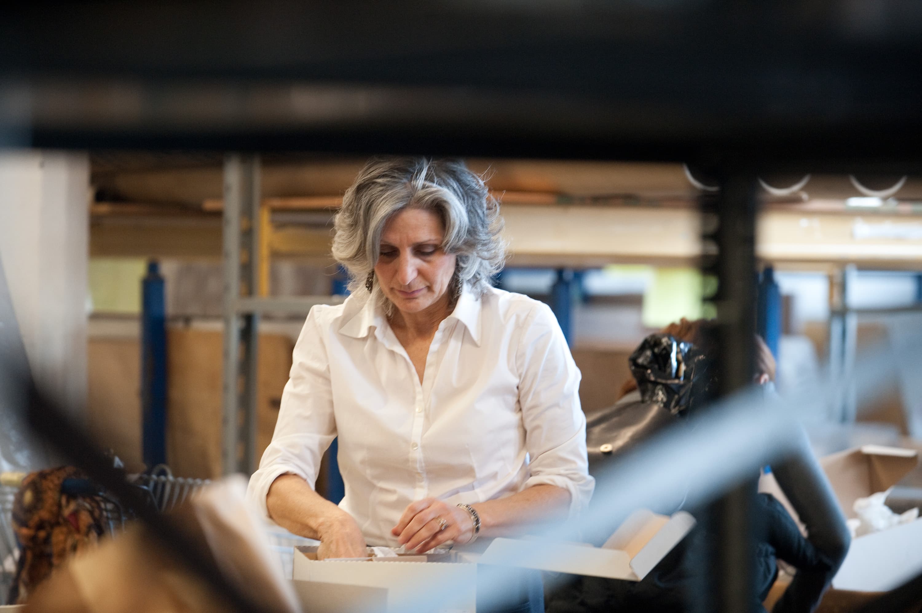 A woman with gray hair in a white shirt working in a workshop, surrounded by shelves and boxes.