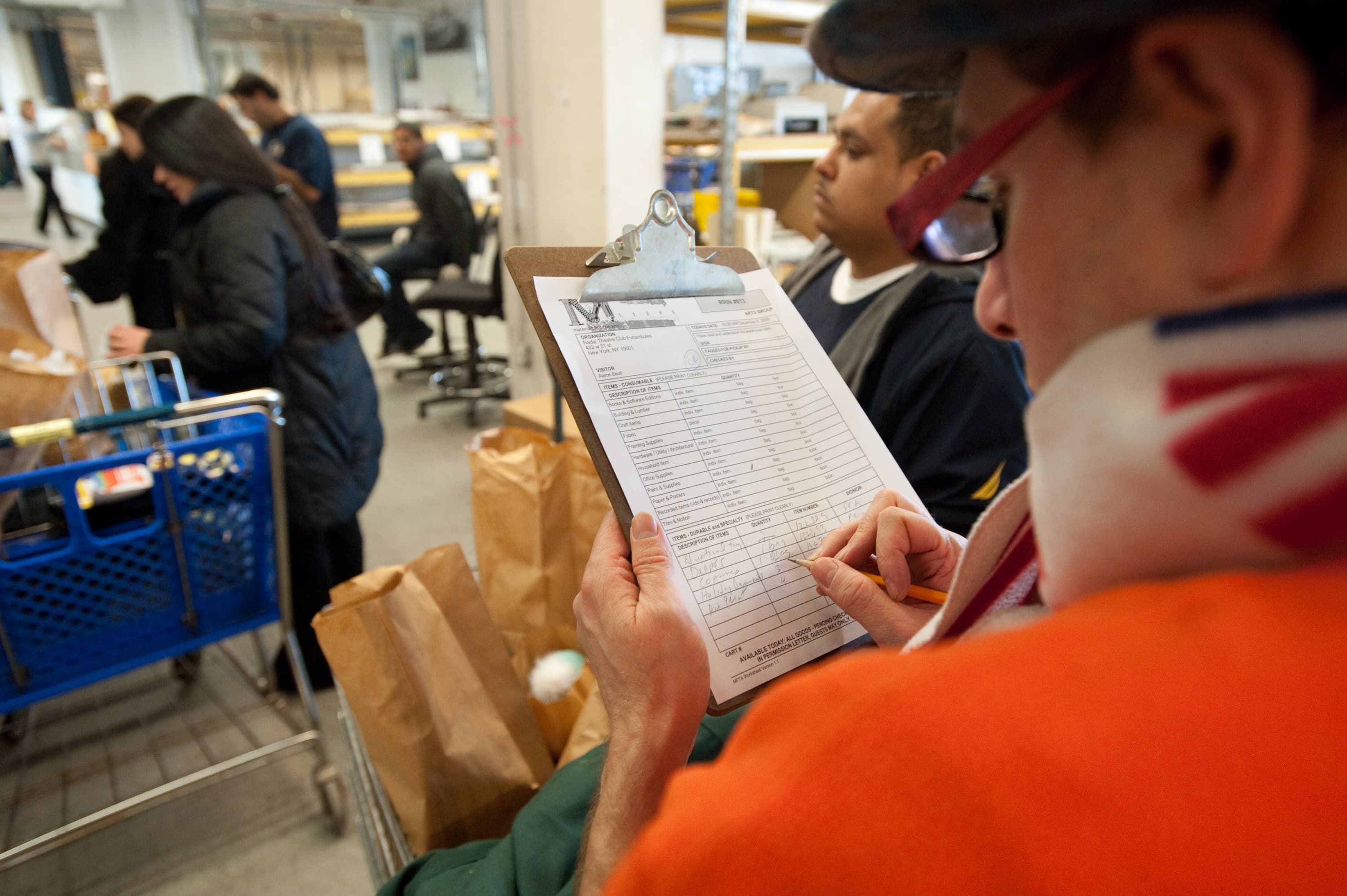 A person writing on a clipboard in a room with people and grocery bags. The clipboard has a form labeled 'M' with various categories filled out. A shopping cart and several brown paper bags are in the background.