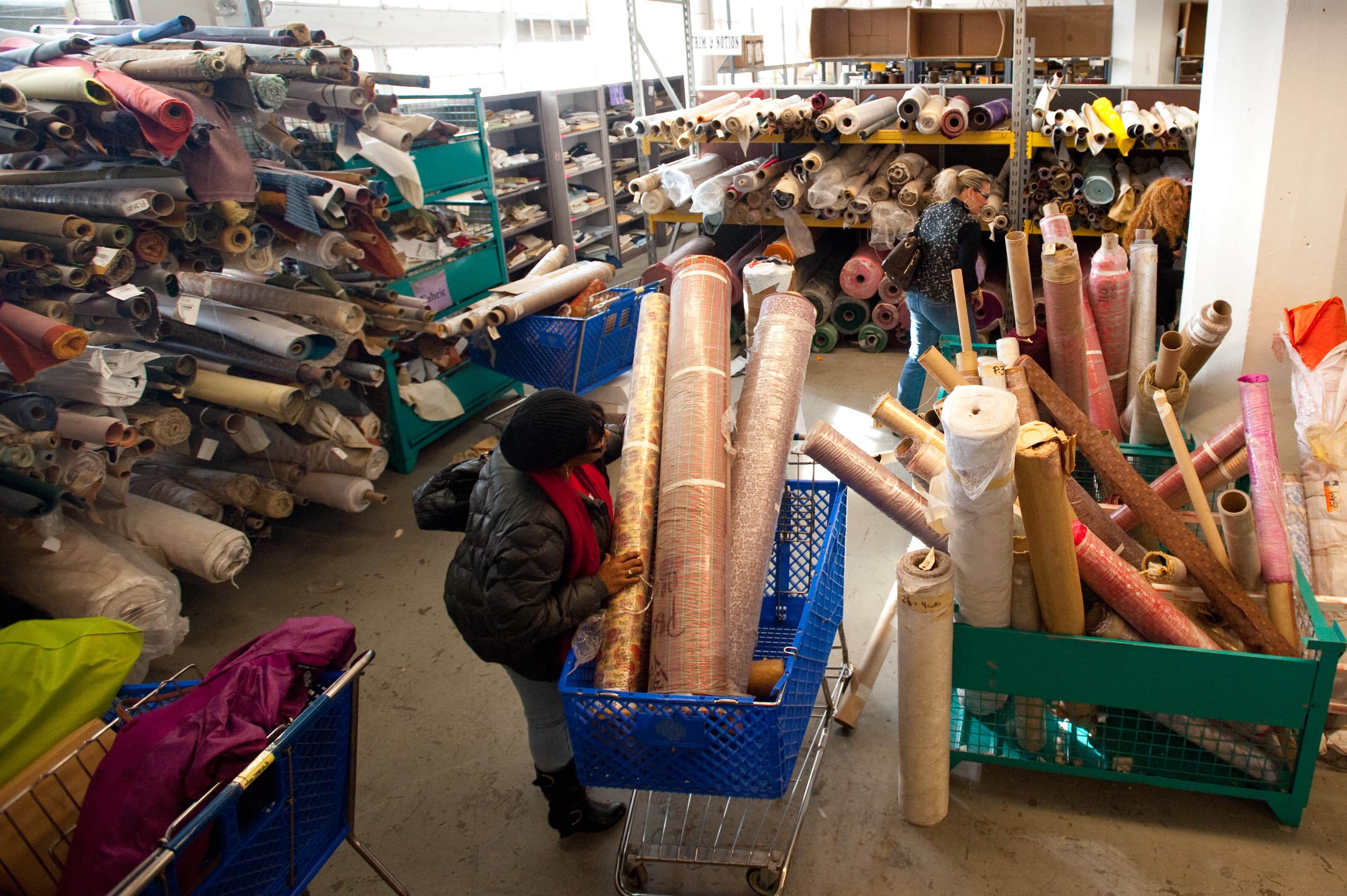 A fabric store with numerous rolls of fabric on shelves and in shopping carts. Several people are browsing through the materials.