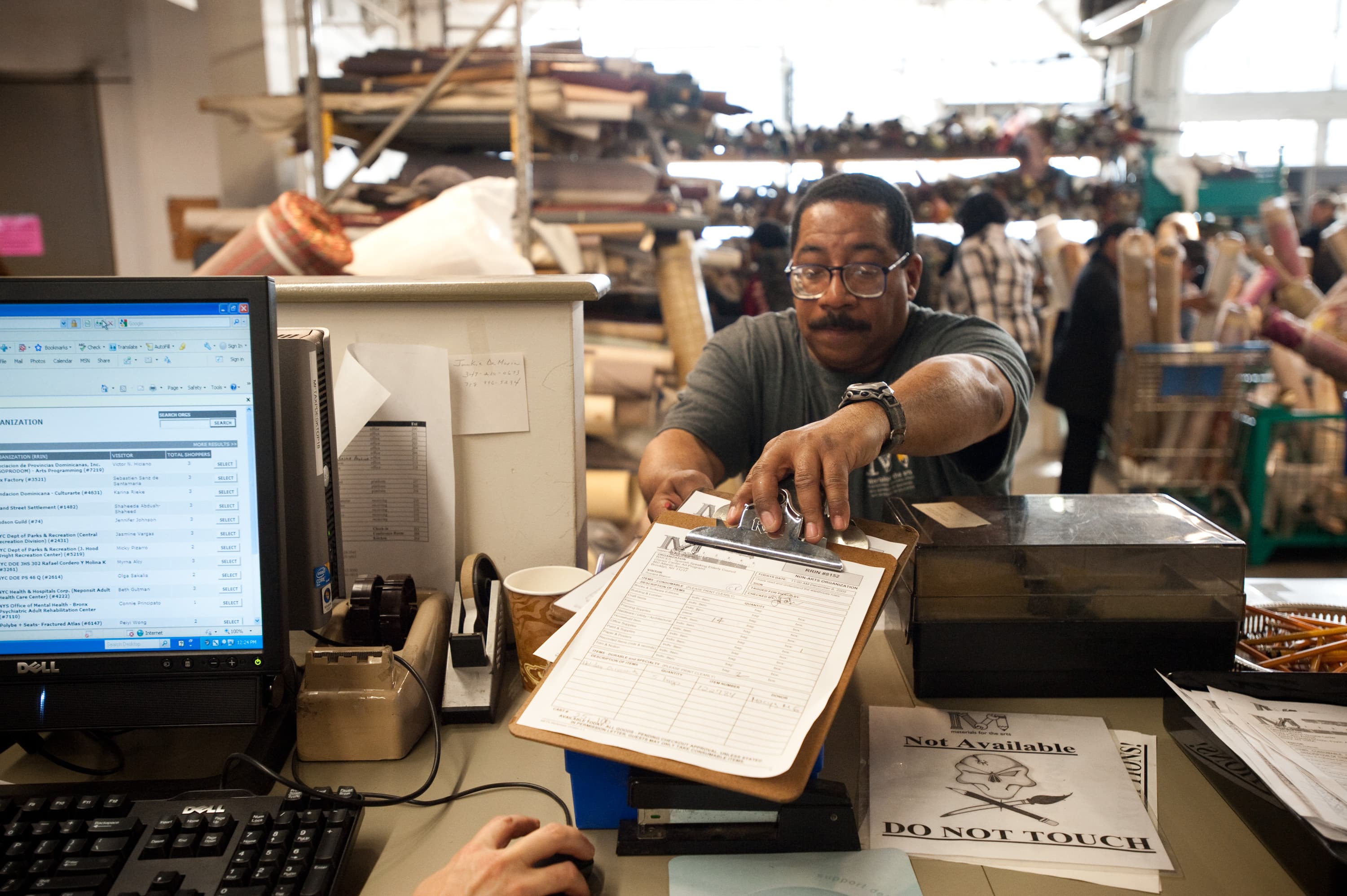 A person reaching across a counter to hand over a clipboard with paperwork. A computer screen displays a list, and there are various office supplies, including a keyboard, tape dispenser, and papers with a 'DO NOT TOUCH' sign, on the desk. In the background, people and rolled fabrics are visible in a warehouse setting.