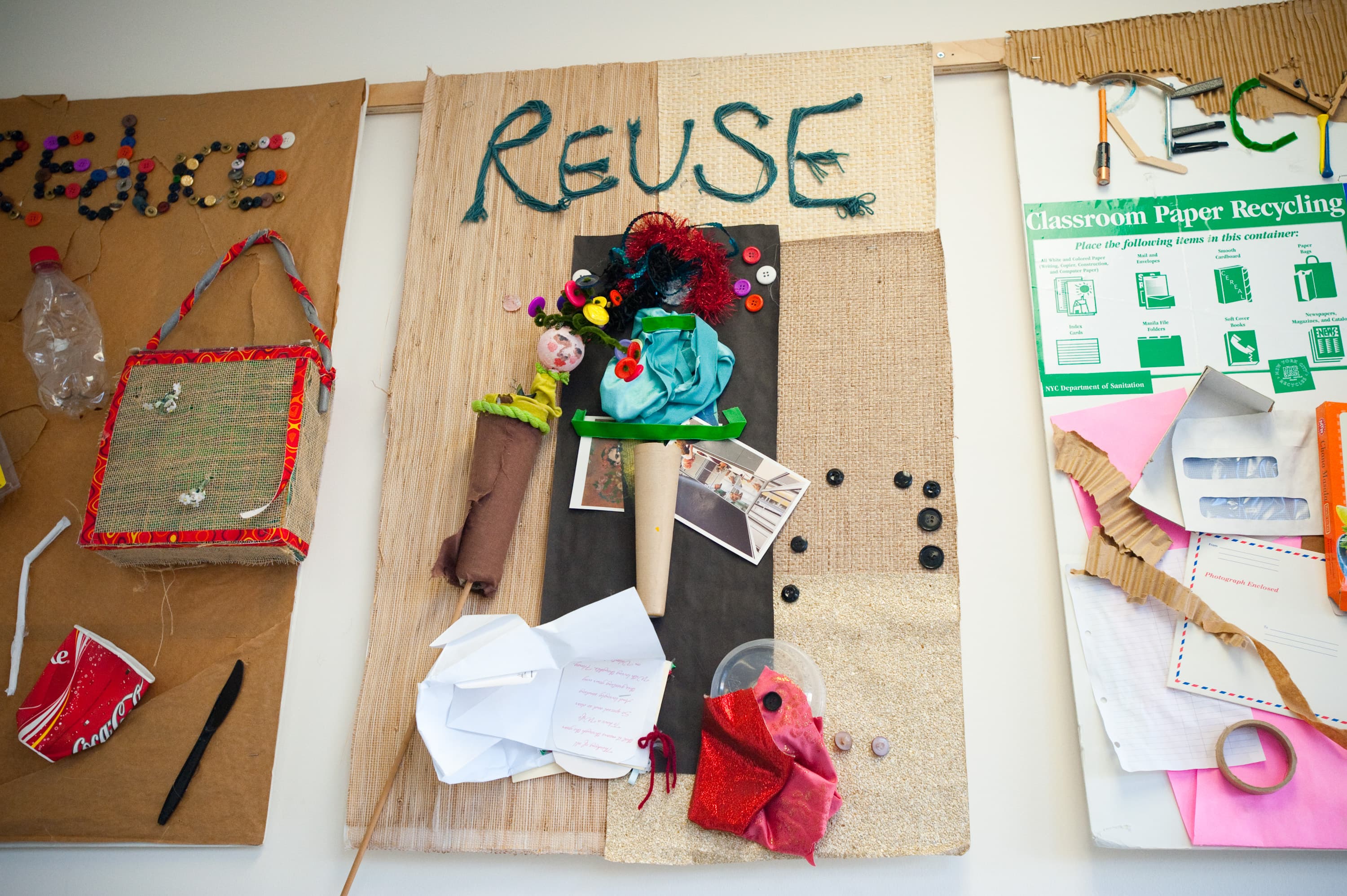 Various art pieces related to recycling. There is a panel labeled "REUSE" with assorted materials such as buttons, fabric, and photographs. Another panel has a classroom paper recycling sign with instructions from the NYC Department of Sanitation, surrounded by paper and cardboard scraps.