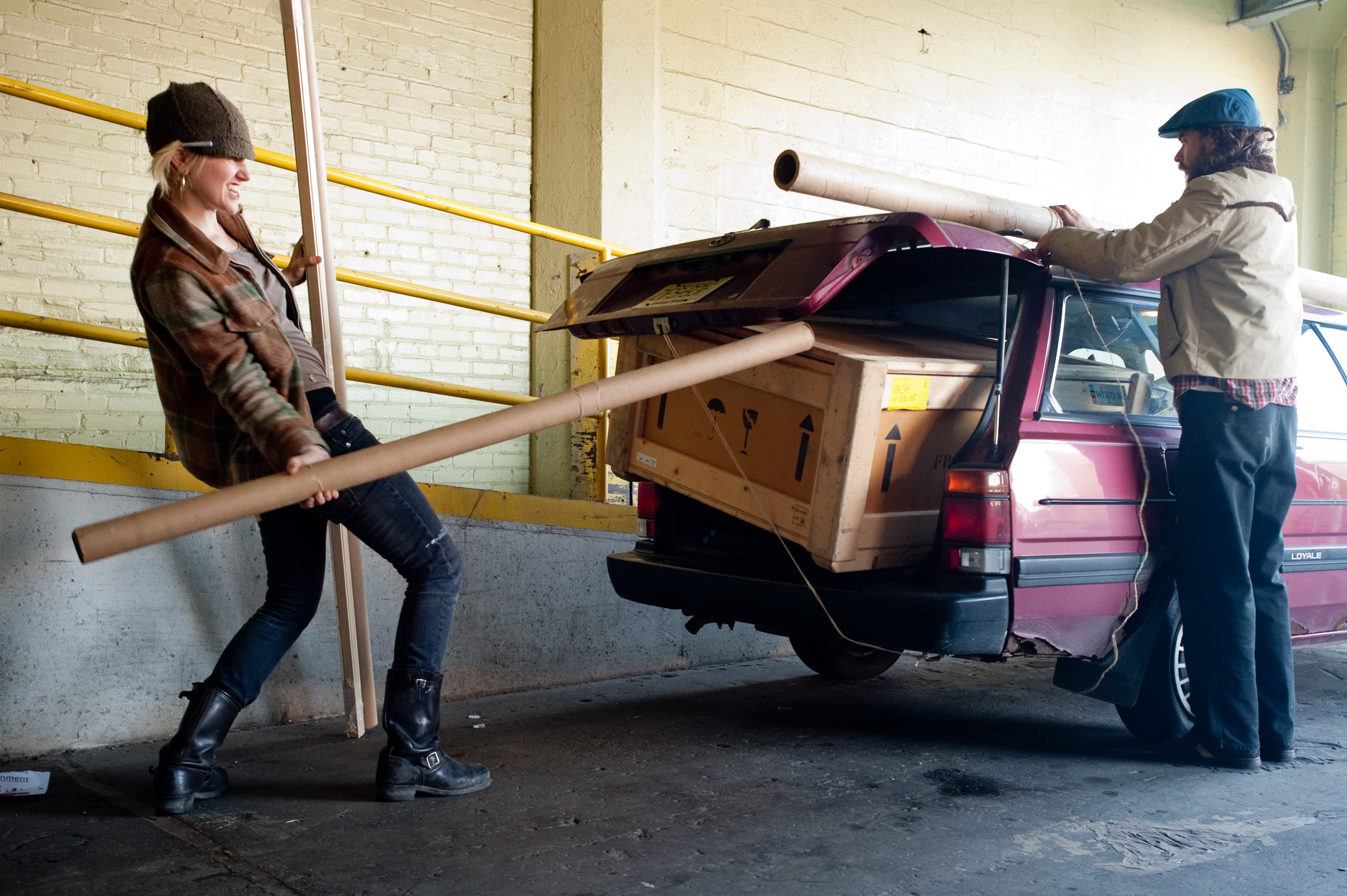 Two people loading long cardboard tubes and a wooden crate into the back of a red station wagon in a warehouse setting.