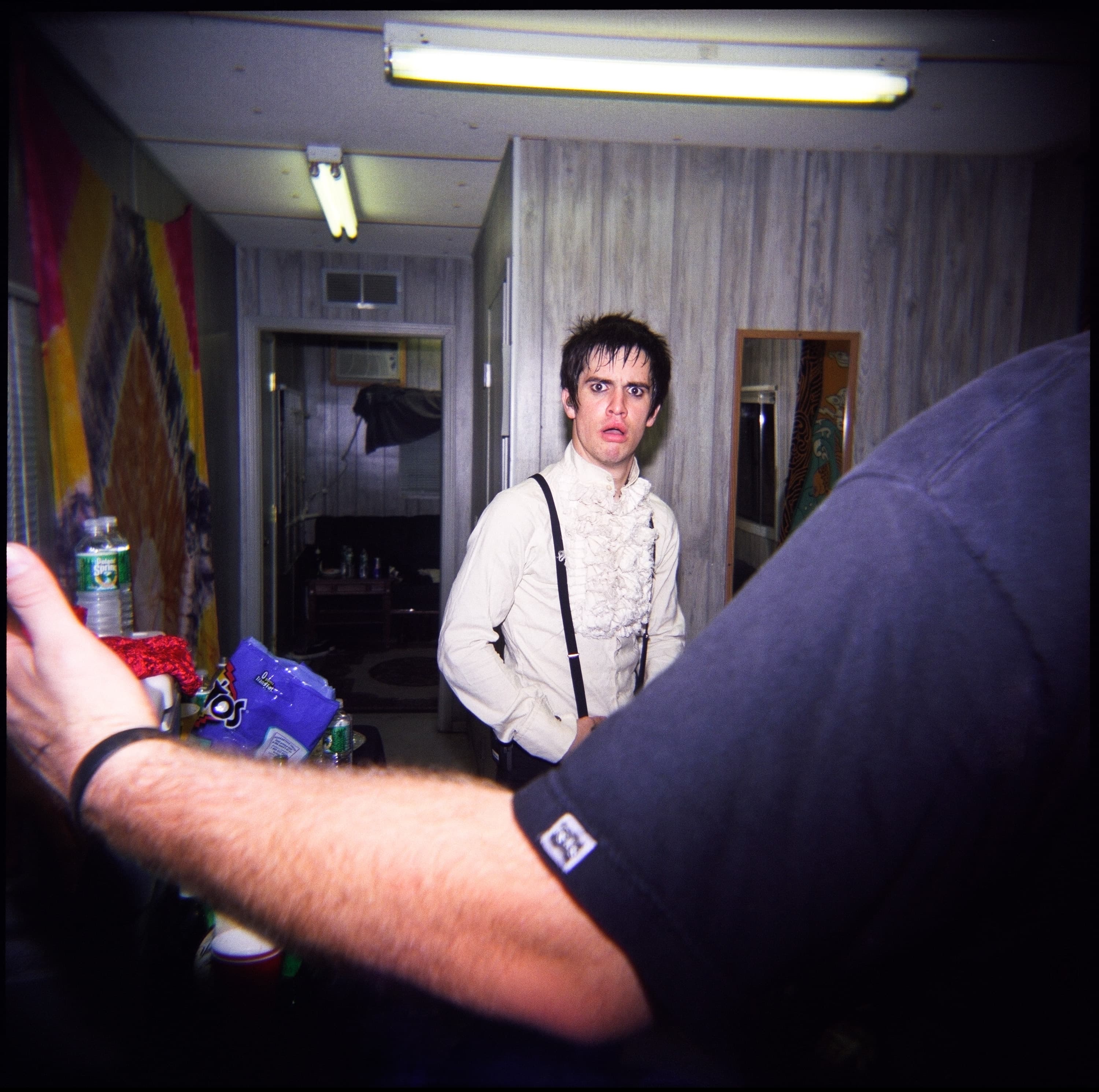 A person wearing a ruffled shirt and suspenders stands in a room, looking towards the camera. A person's arm is seen in the foreground reaching out. The room has a decorated wall with a colorful pattern, a mirror, and various items on a table, including a water bottle and snack bags.