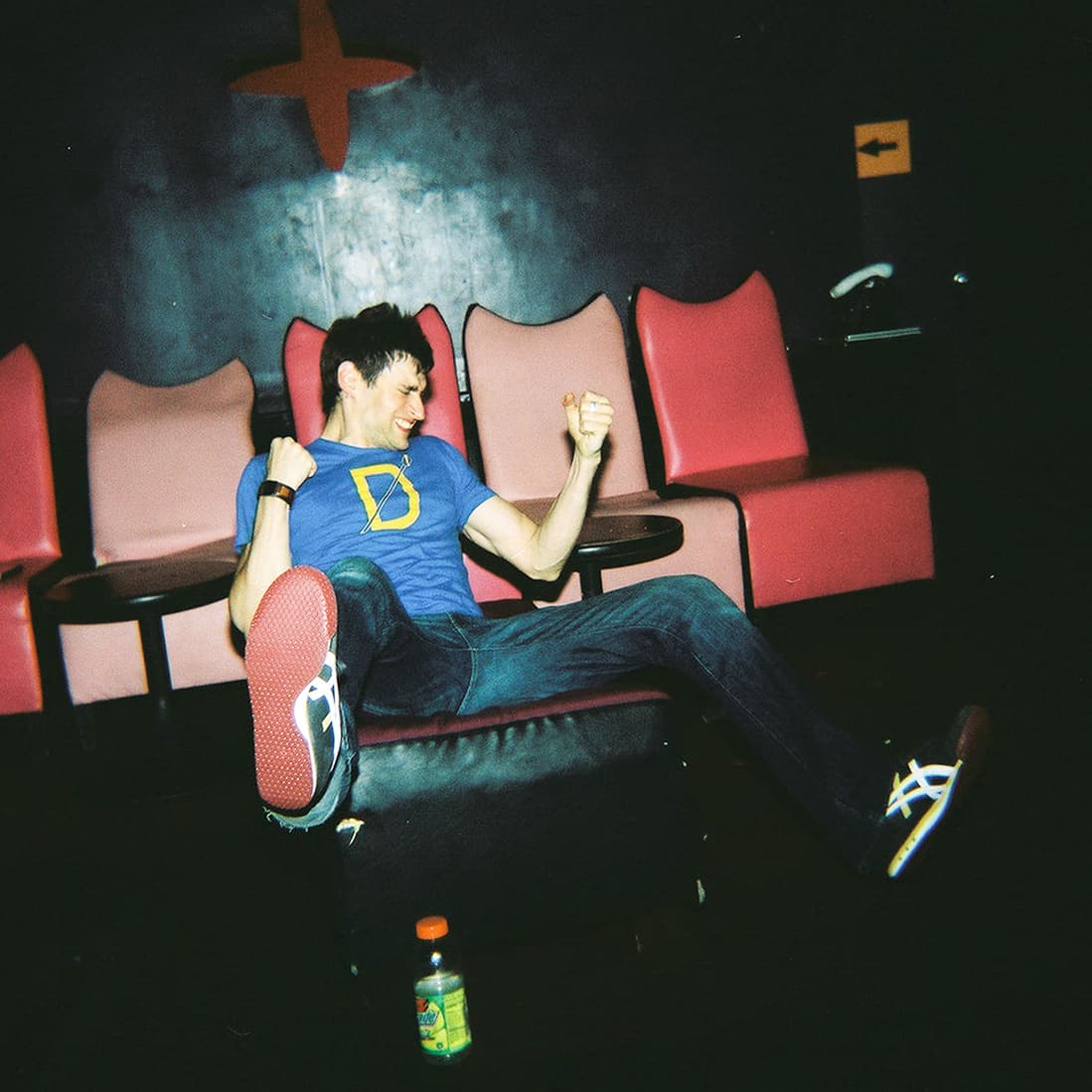 A person wearing a blue shirt with a yellow design, sitting on a black object, playfully raising their fists. There are pink cushioned chairs and a small table in the background. A bottle is on the floor.