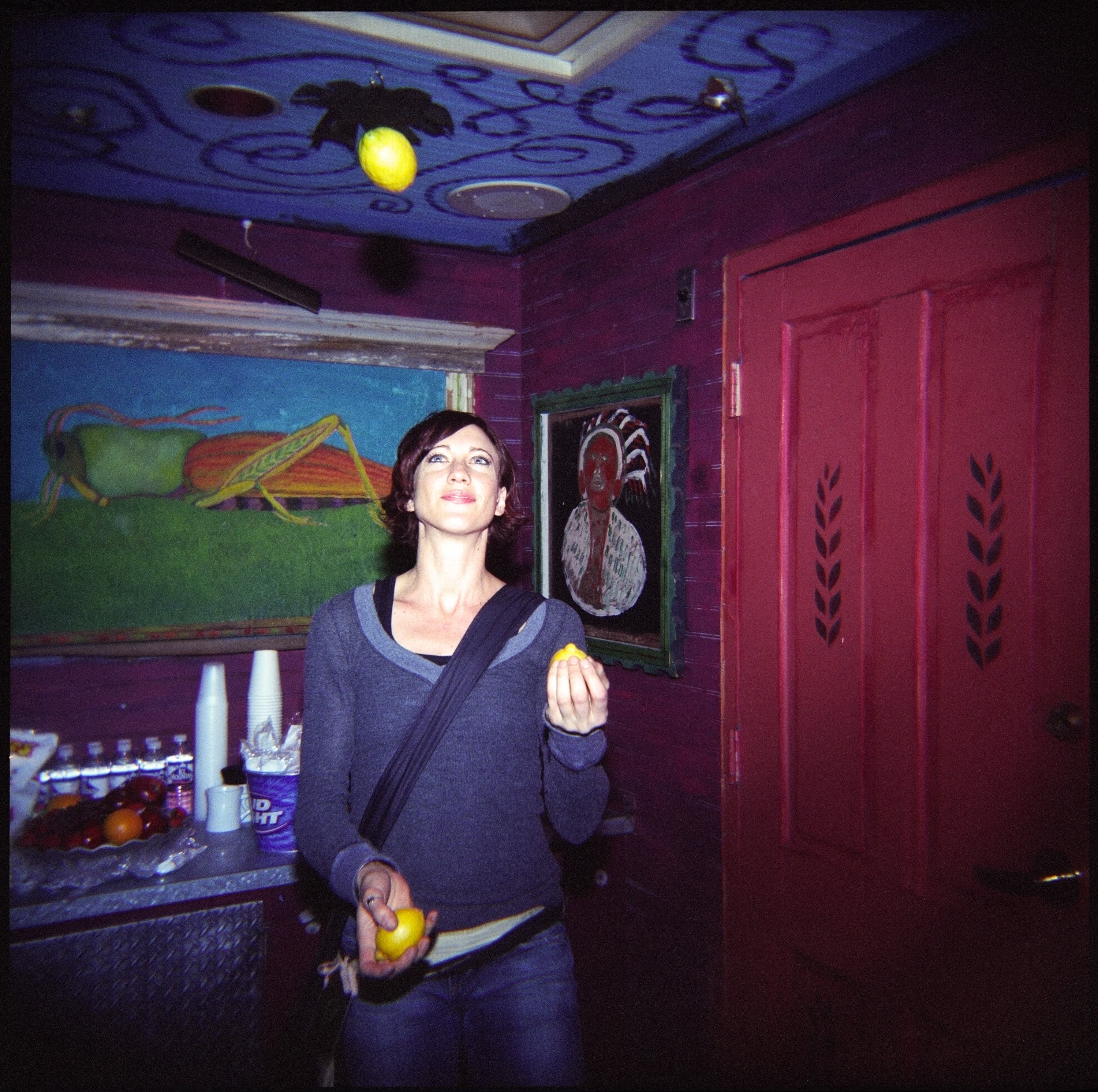 A person is standing indoors juggling lemons. The room has a painted blue ceiling with swirls, red walls, and two framed artworks. A counter with bottles and a string of cups is in the background.