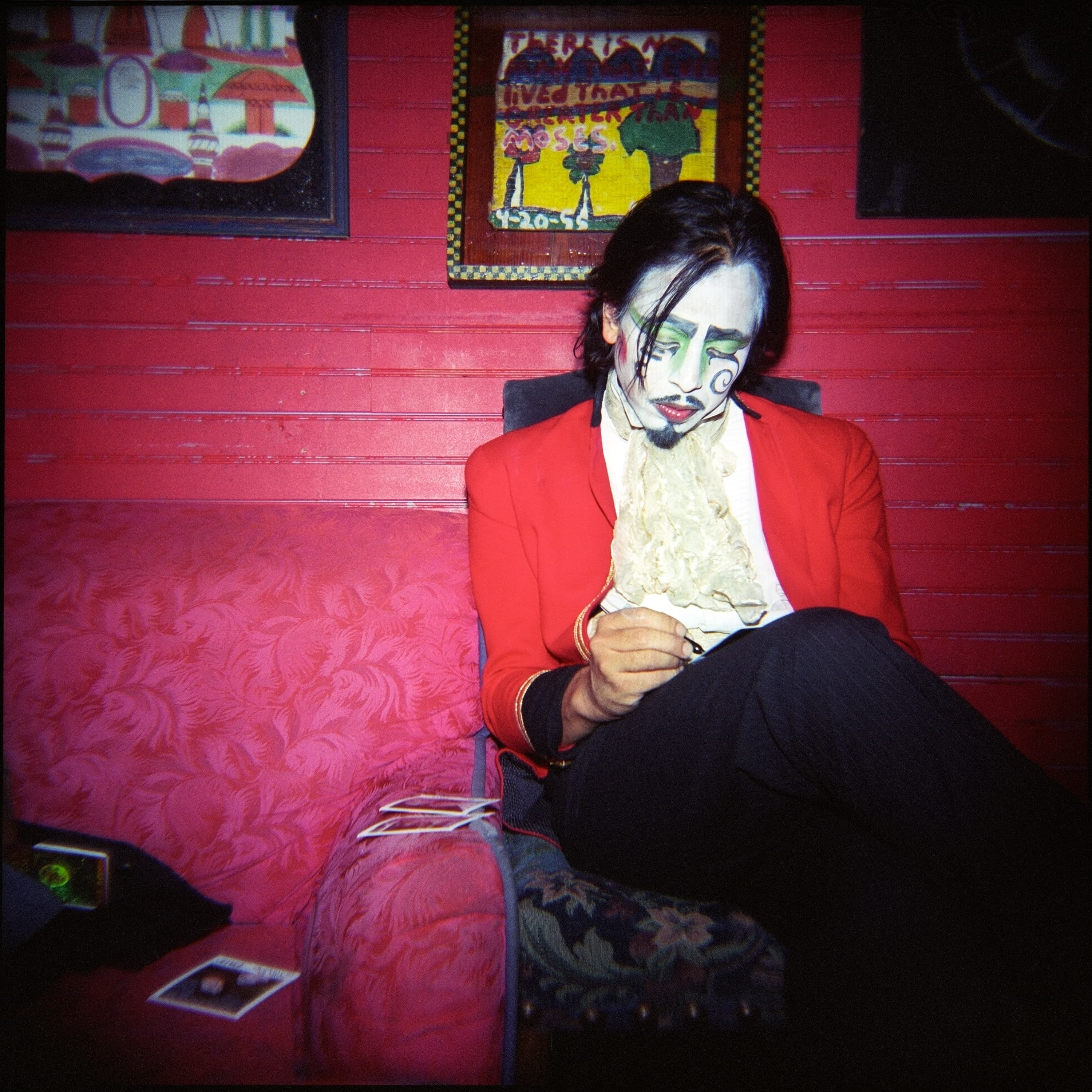 Person with face paint and a red blazer sitting on a pink-patterned couch, holding a small object. The background has red paneling and artworks on the wall.