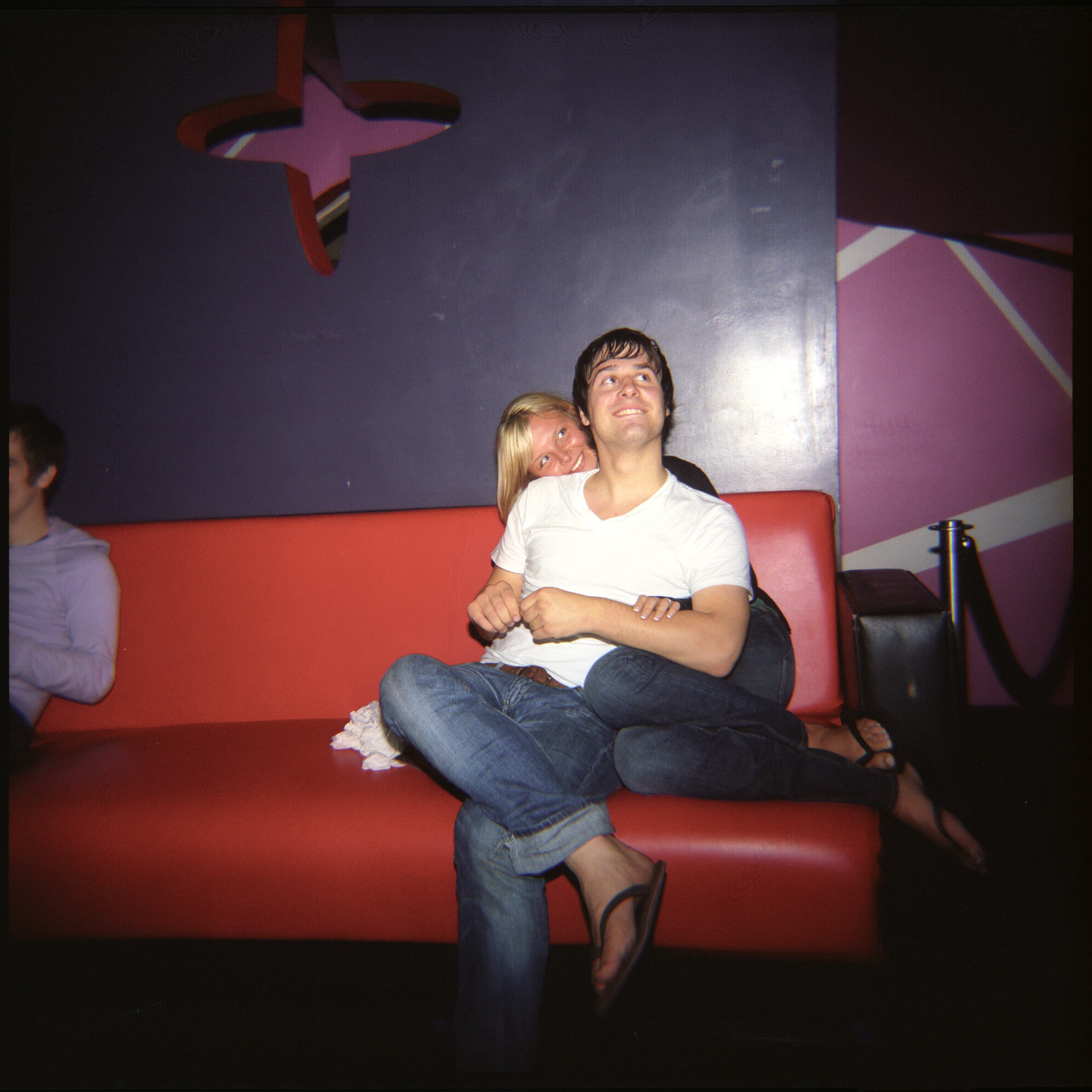 Two people sitting on a red couch, with one person hugging the other from behind. There is a decorative element on the wall in the shape of a star. A third person is partially visible on the left side of the image.