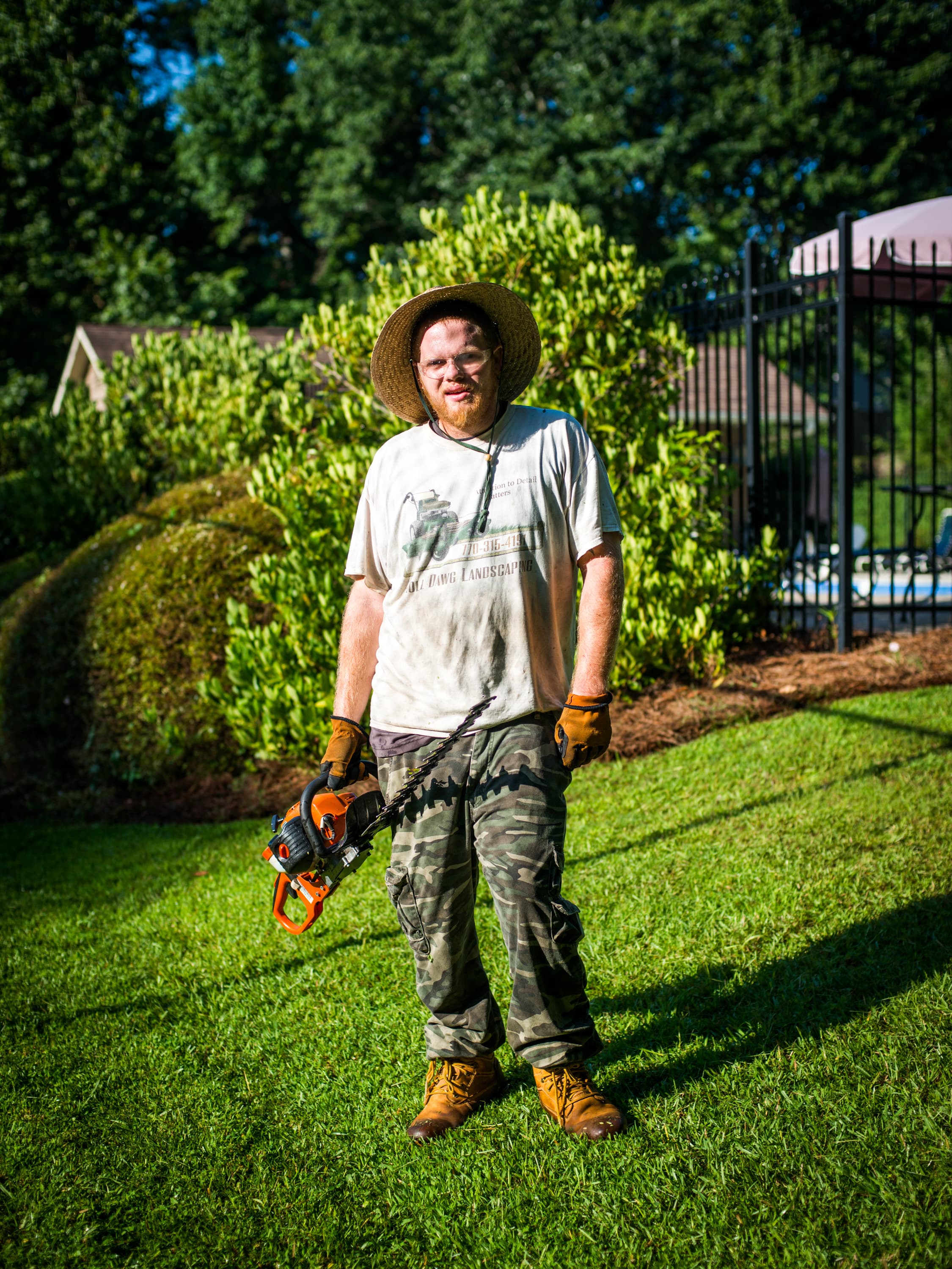 Person standing on a lawn wearing a straw hat, camo pants, and boots, holding a hedge trimmer. There are bushes and a black metal fence in the background.