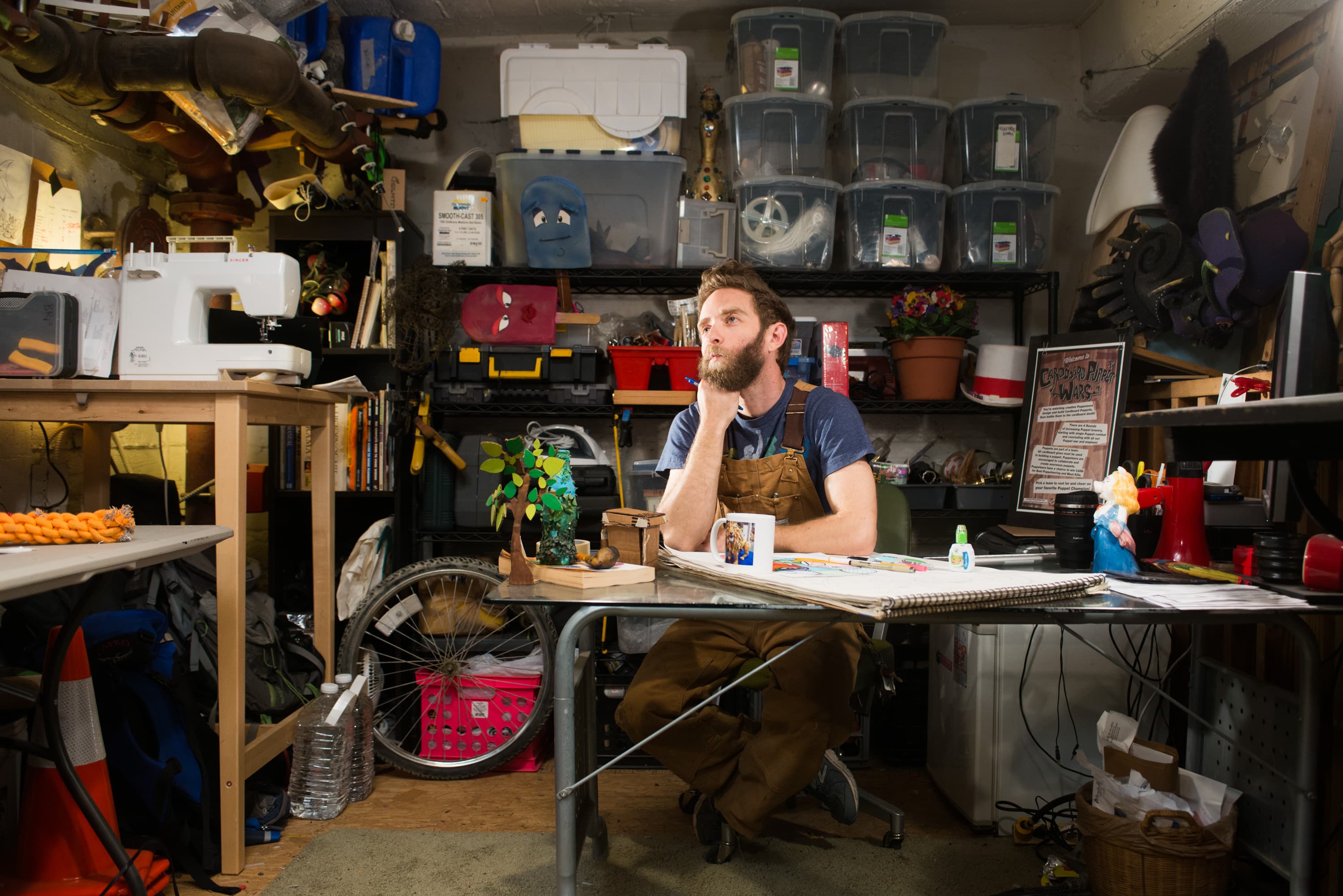 A person in a workshop filled with various items. They sit at a table with a mug and a small model tree, with shelves of plastic bins and tools behind them. A sewing machine and other crafting materials are visible around the room.