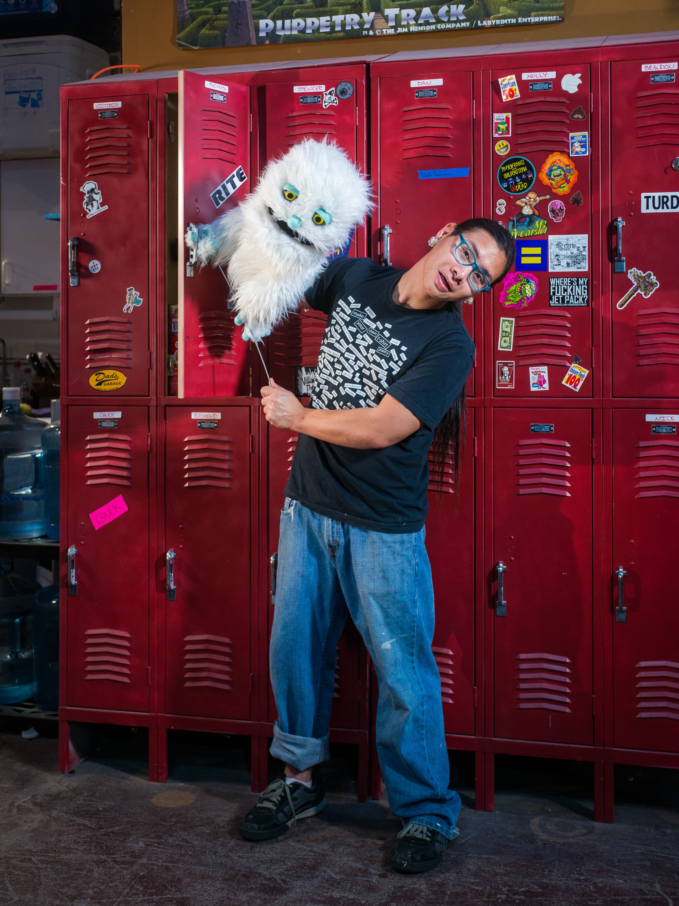 A person holding a white furry puppet in front of a row of red lockers decorated with various stickers and labels.