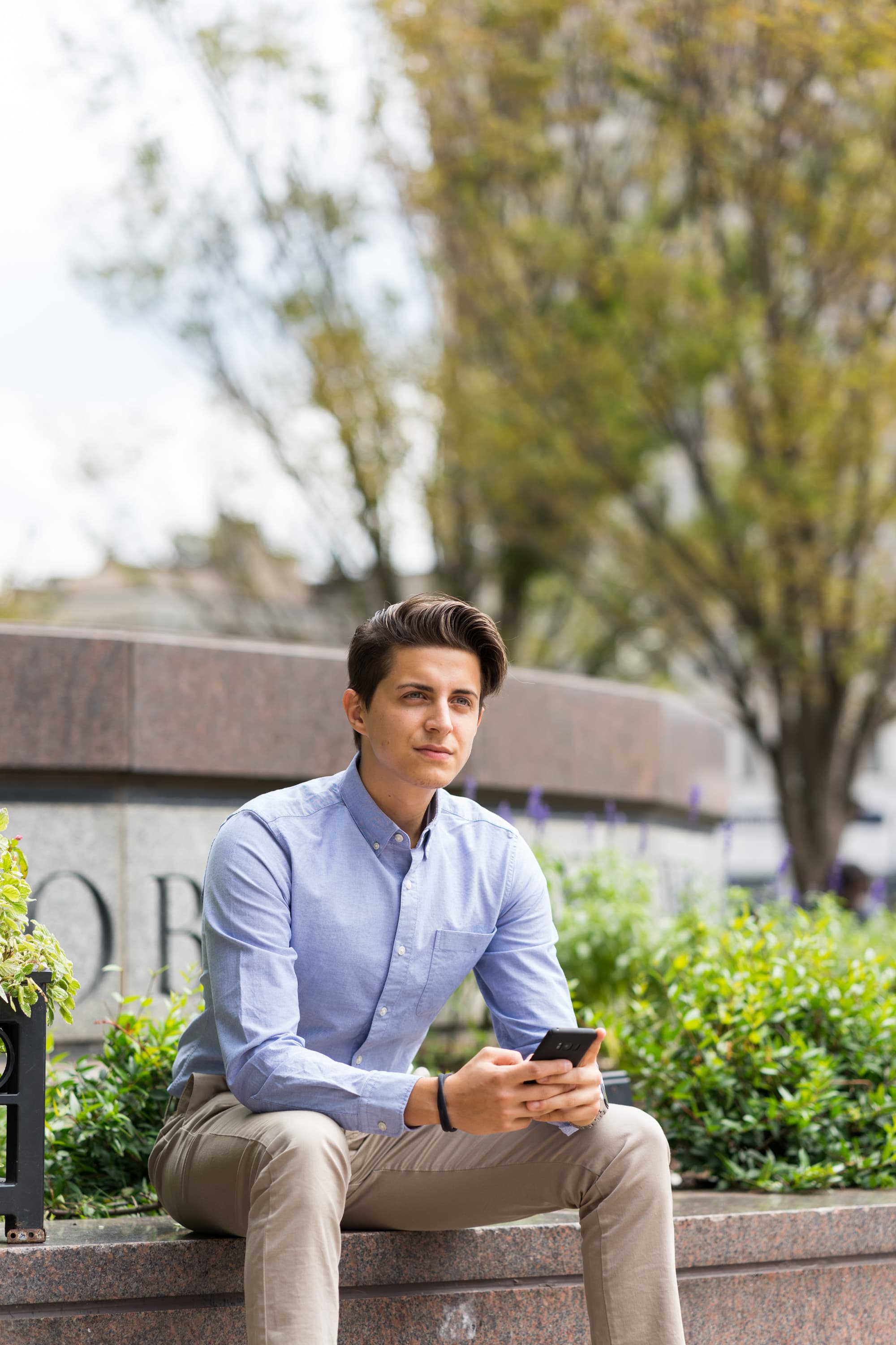 A person sitting on a stone ledge outdoors, holding a smartphone. They are wearing a light blue shirt and beige pants. Surrounding the ledge are green plants and trees are visible in the background.