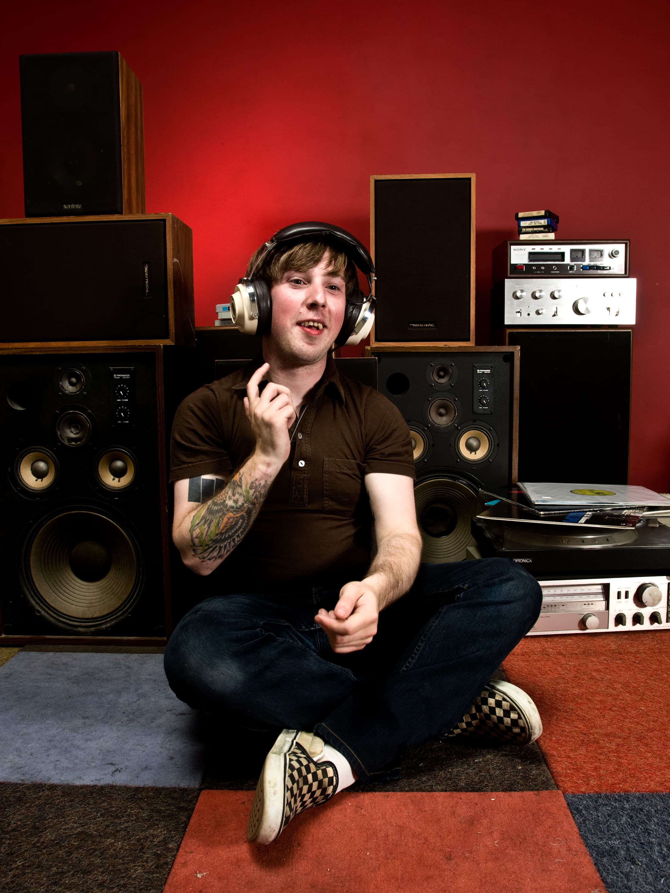 A person sitting on a colorful carpet wearing headphones, surrounded by vintage audio equipment including speakers, a turntable, and a stereo receiver. They have a tattoo on one arm and are wearing a brown shirt and checkered shoes.