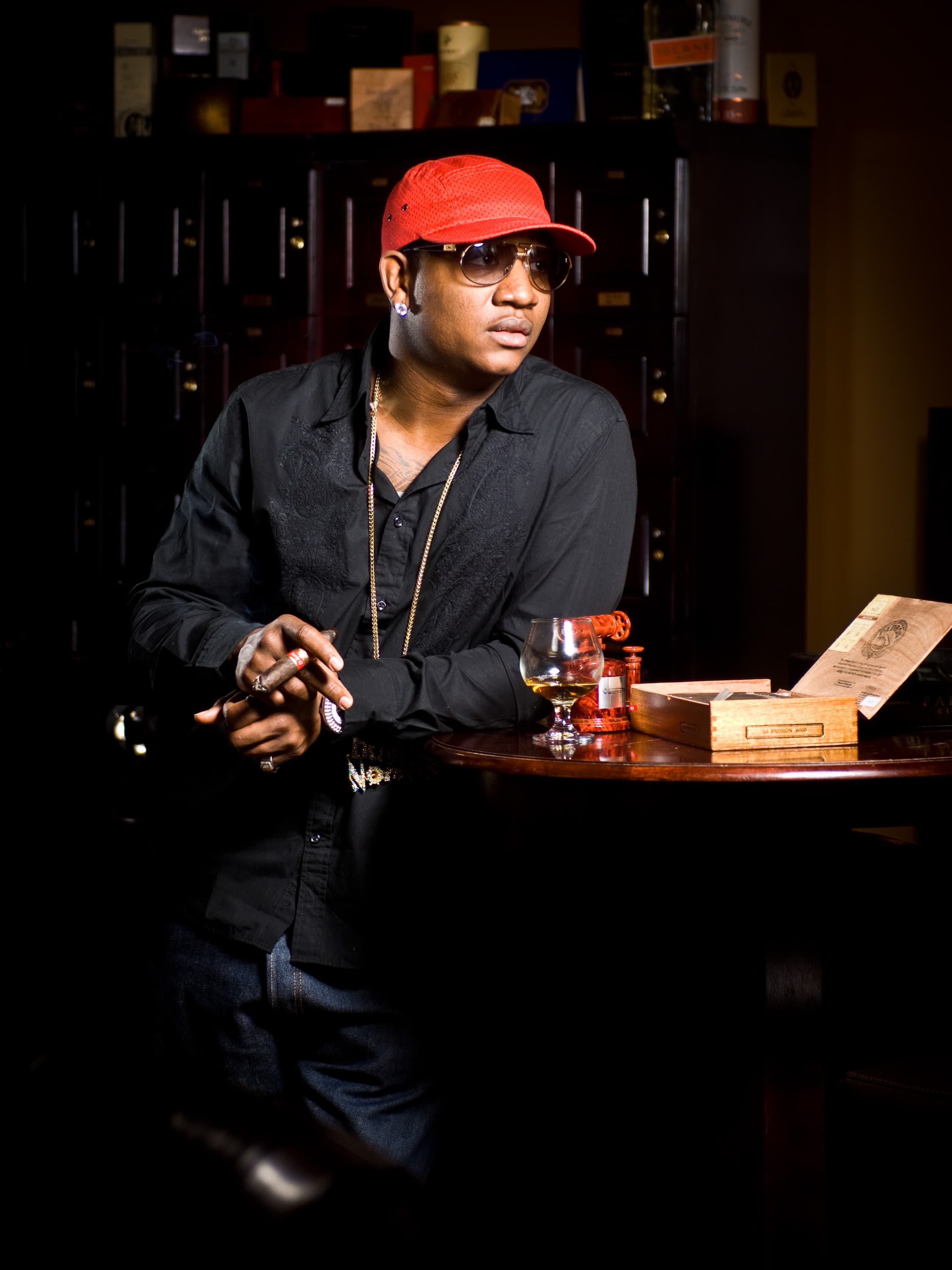 Rapper Yung Joc wearing a red cap, sunglasses, and a black shirt stands next to a table with a glass of drink and an open cigar box. They are holding a cigar and wearing a gold chain. The background features shelves with various items.