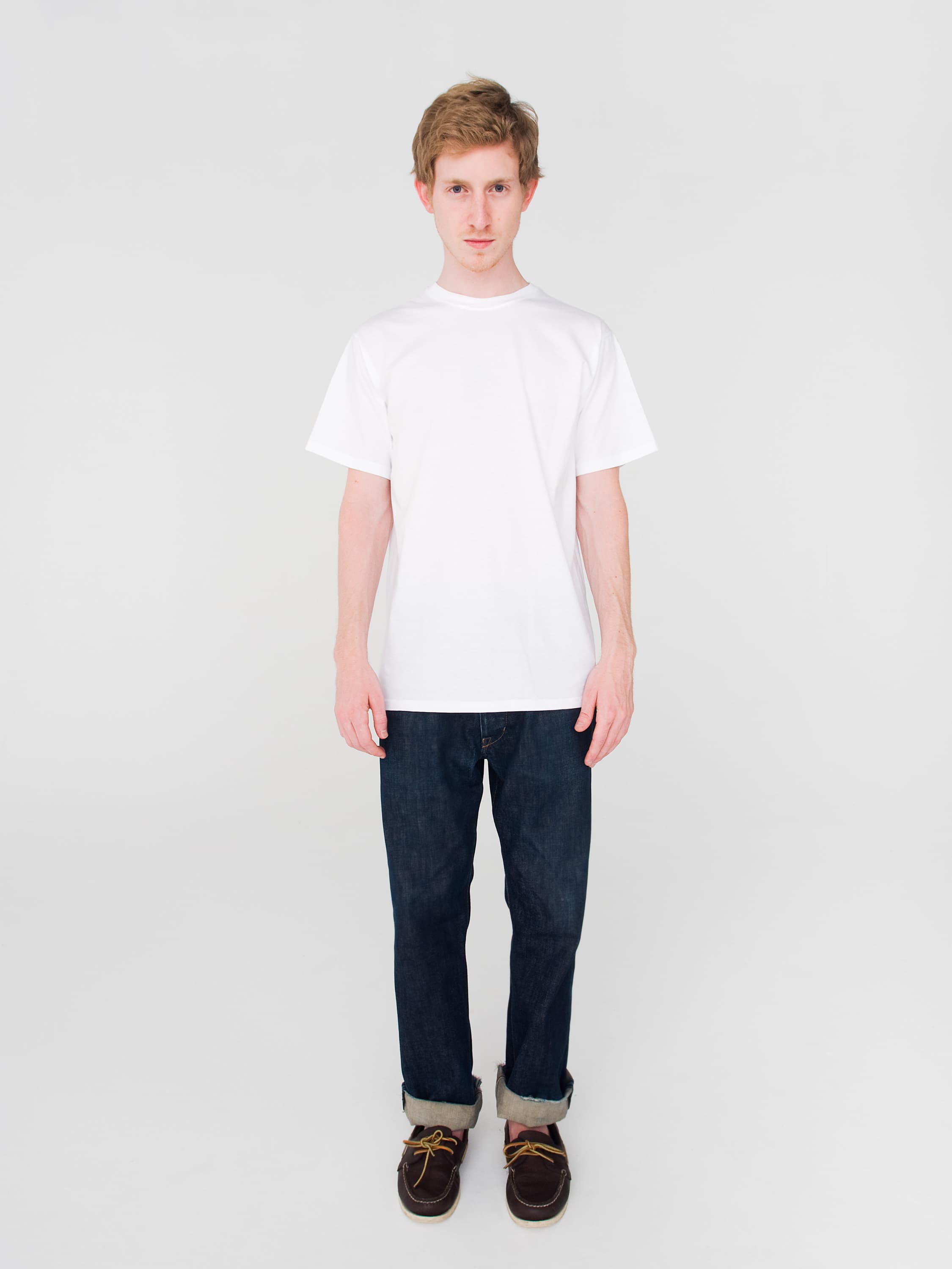 Rapper Asher Roth wearing a plain white T-shirt, dark blue jeans, and brown shoes stands against a plain background.