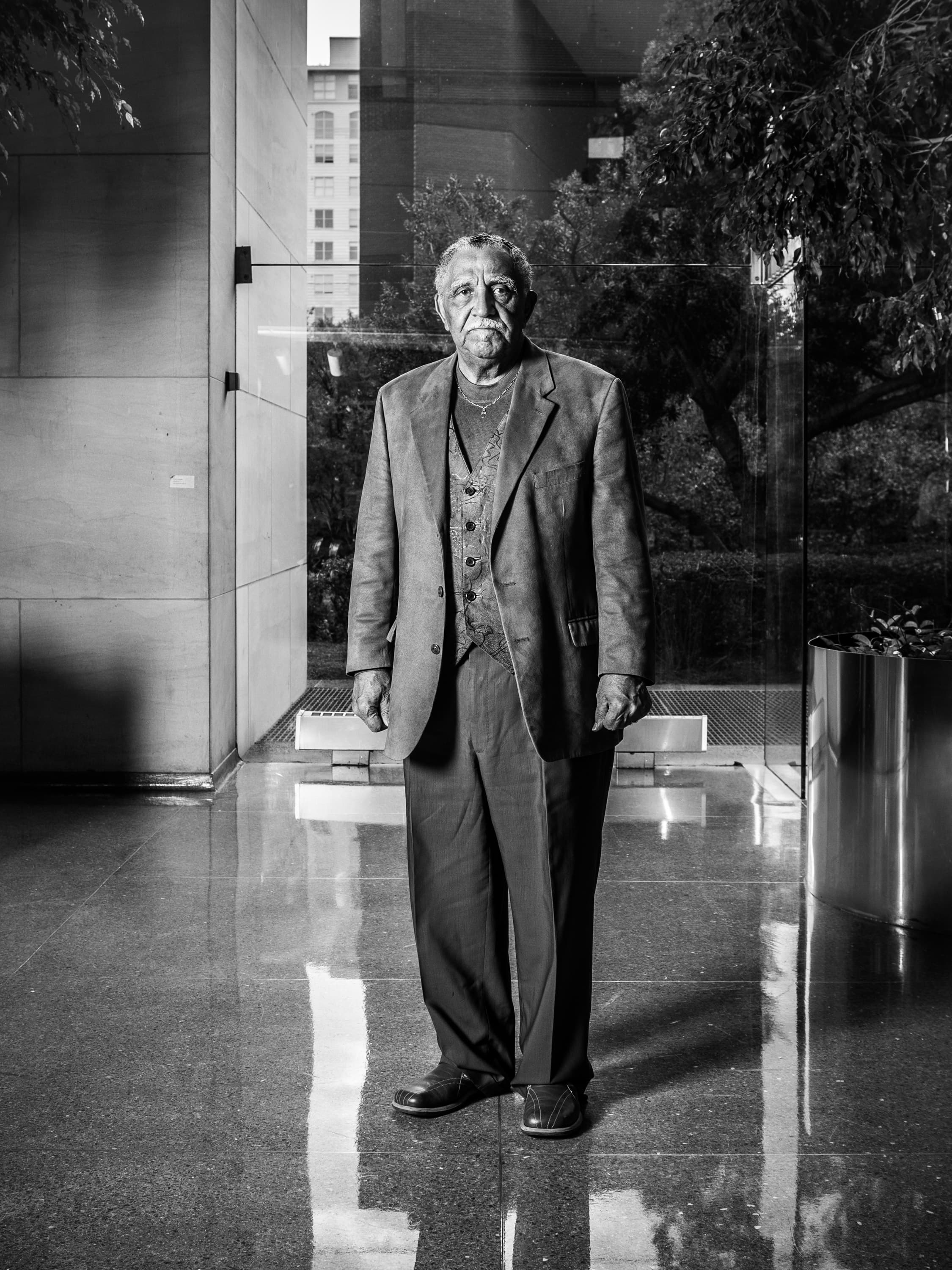 Civil rights leader Joseph Lowery in a suit stands indoors with a reflective floor. The background features a large window with a view of trees and buildings outside.