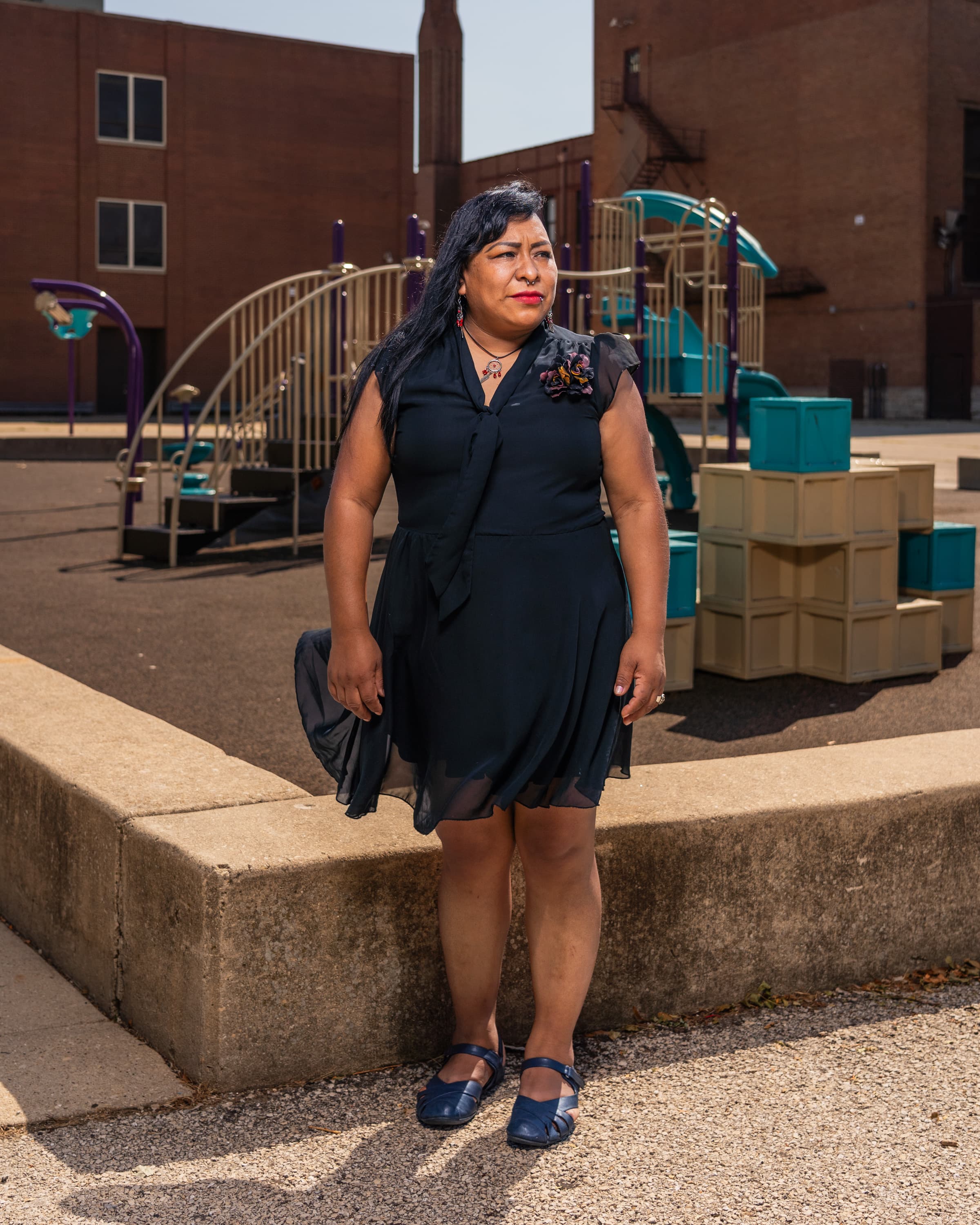 A person standing near a playground, wearing a black dress and blue shoes. The playground equipment is visible in the background along with brick buildings.