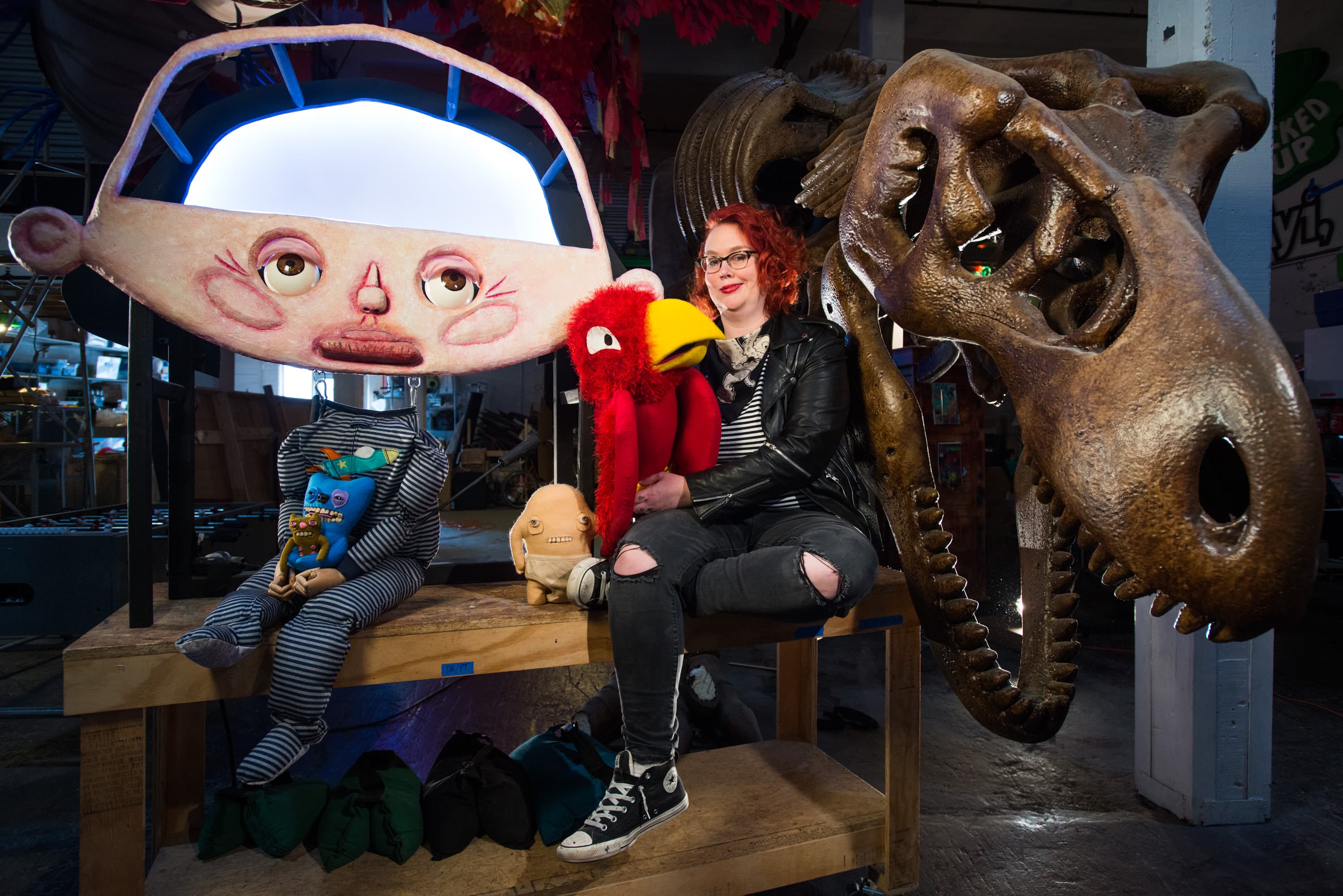 A person with red hair sitting next to a large dinosaur skull, holding a red bird puppet. Nearby, a mannequin with a large, cartoonish face and small dolls sits on a bench.