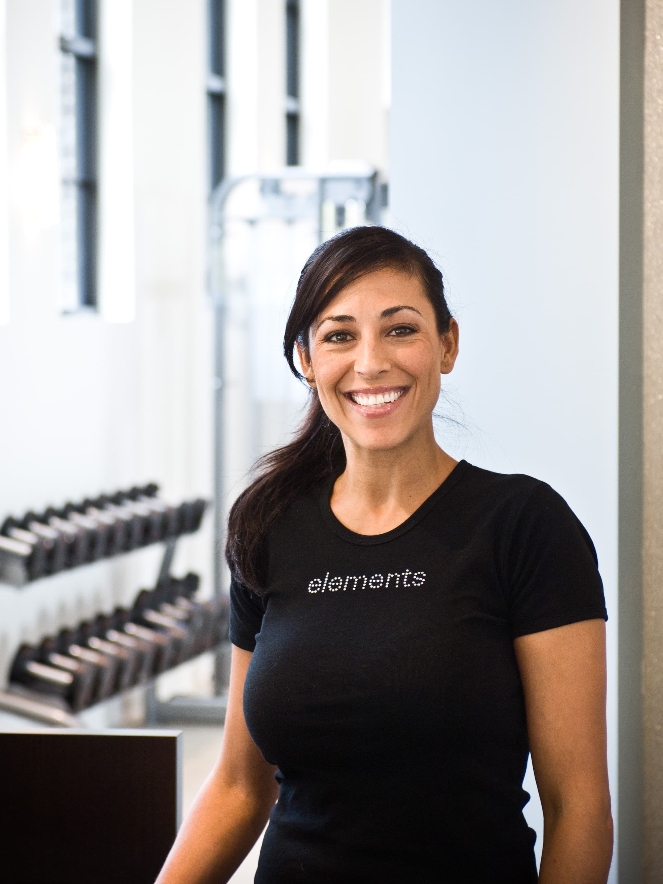 A woman wearing a black t-shirt with the word "elements" on it, standing in a gym with dumbbells and exercise equipment in the background.