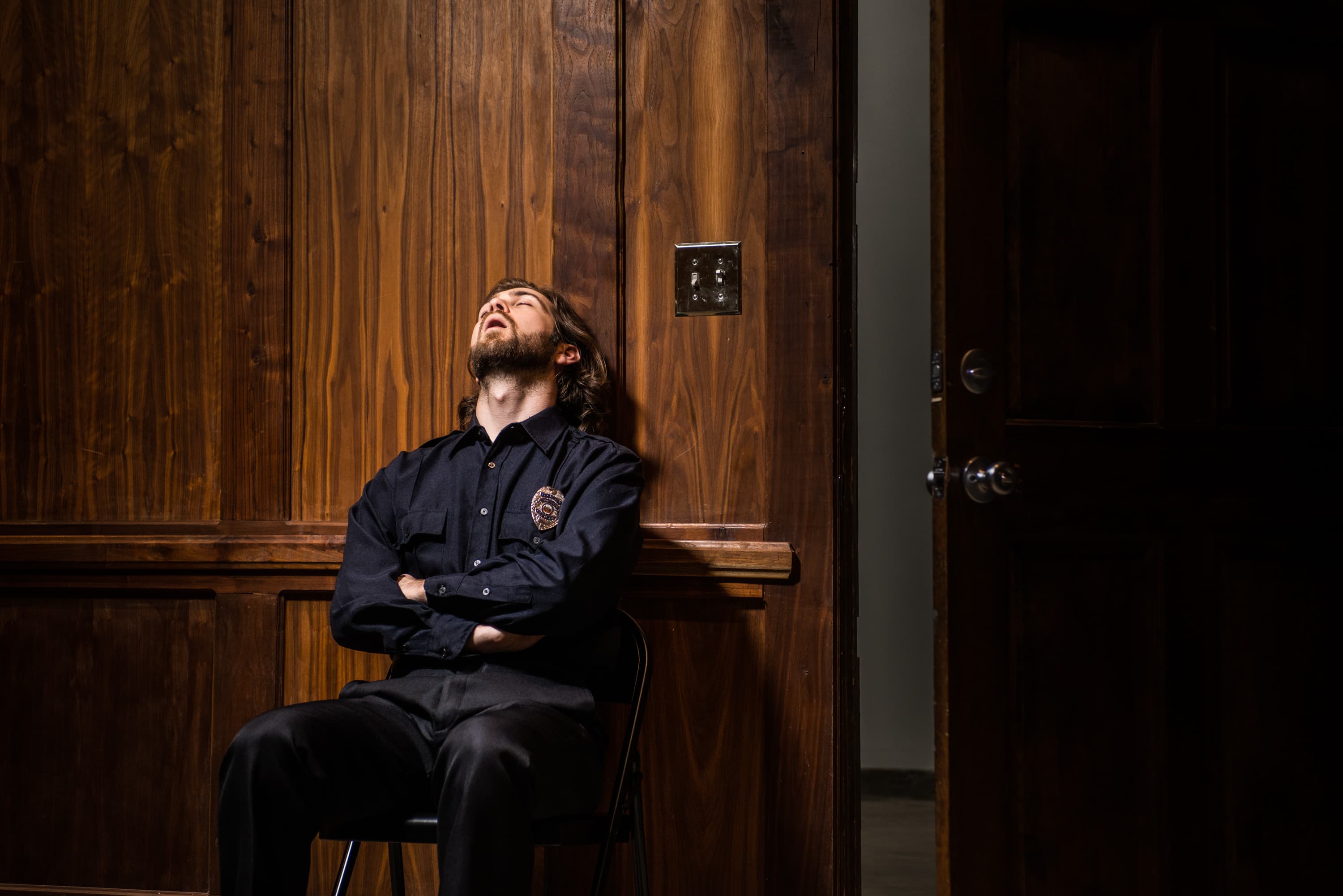 A person in a uniform is sitting on a chair against a wooden wall with their arms crossed and head tilted back, eyes closed. There is a door to the right.