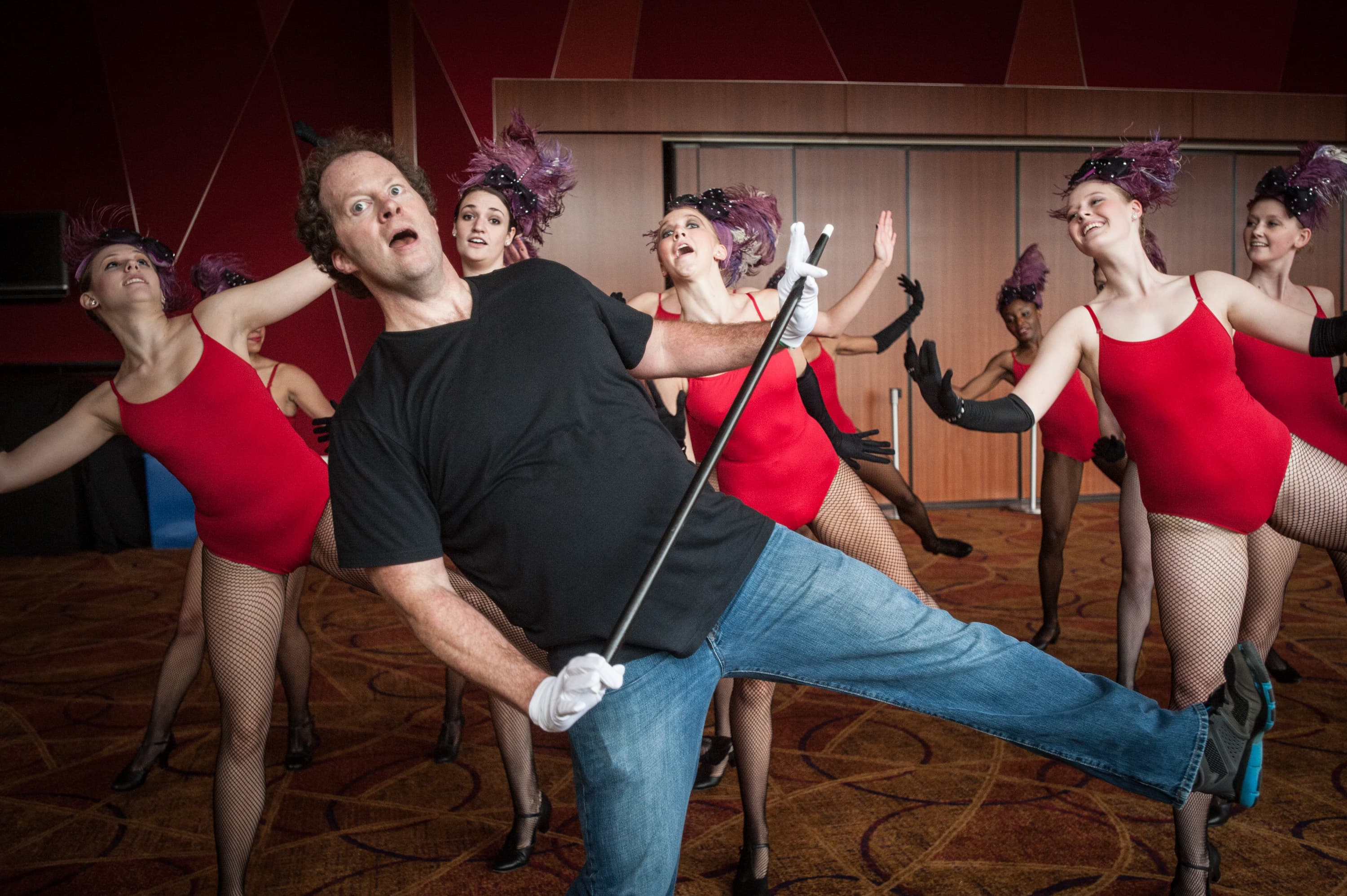 Singer and actor Shuler Hensley in a black shirt and blue jeans holding a cane, surrounded by dancers in red leotards, black gloves, and headpieces, performing in a room with wooden walls.