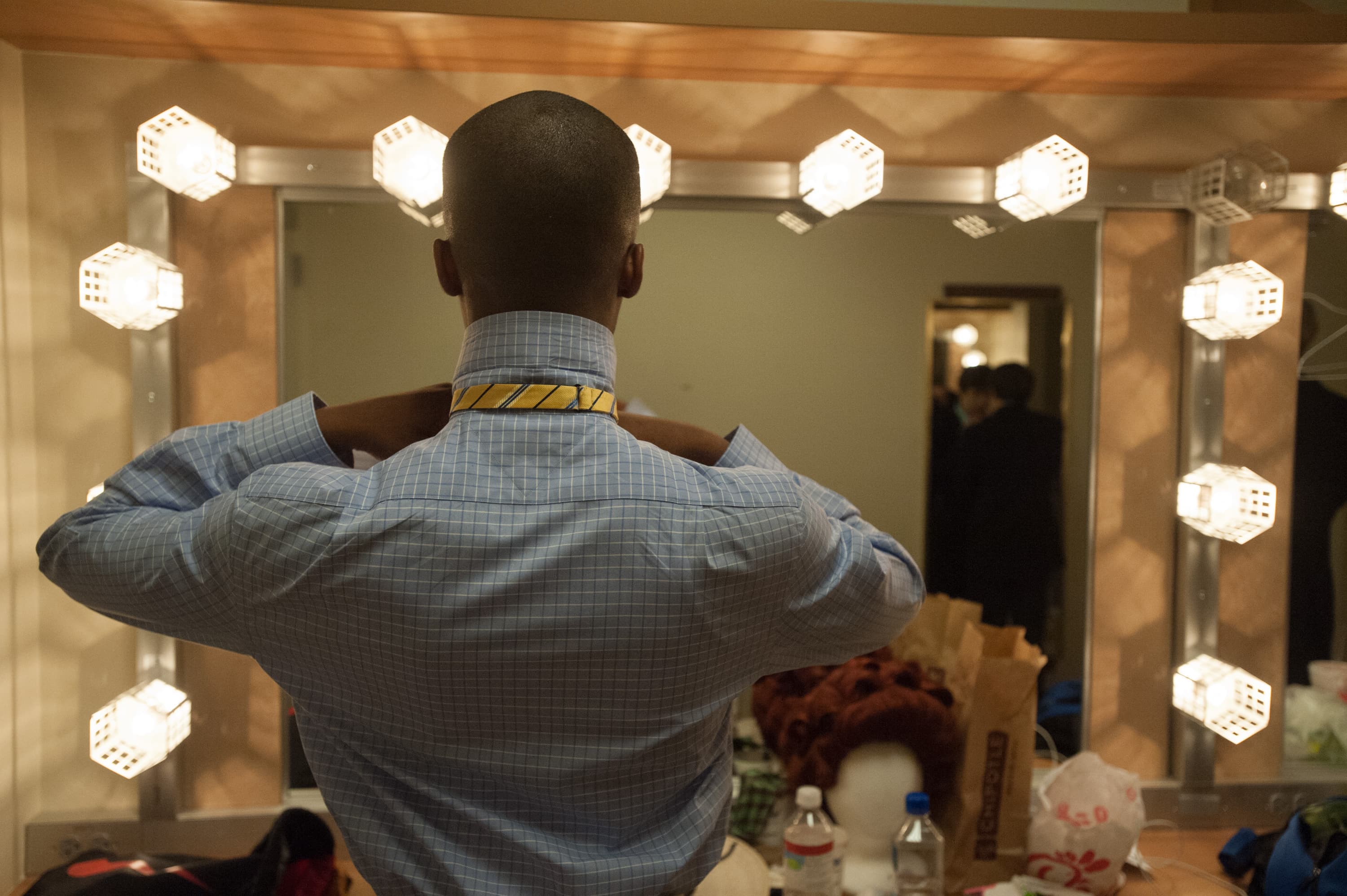 A person adjusting a yellow and black striped necktie in front of a mirror surrounded by lights. The dressing table is cluttered with various items including a wig, paper bags, and water bottles.