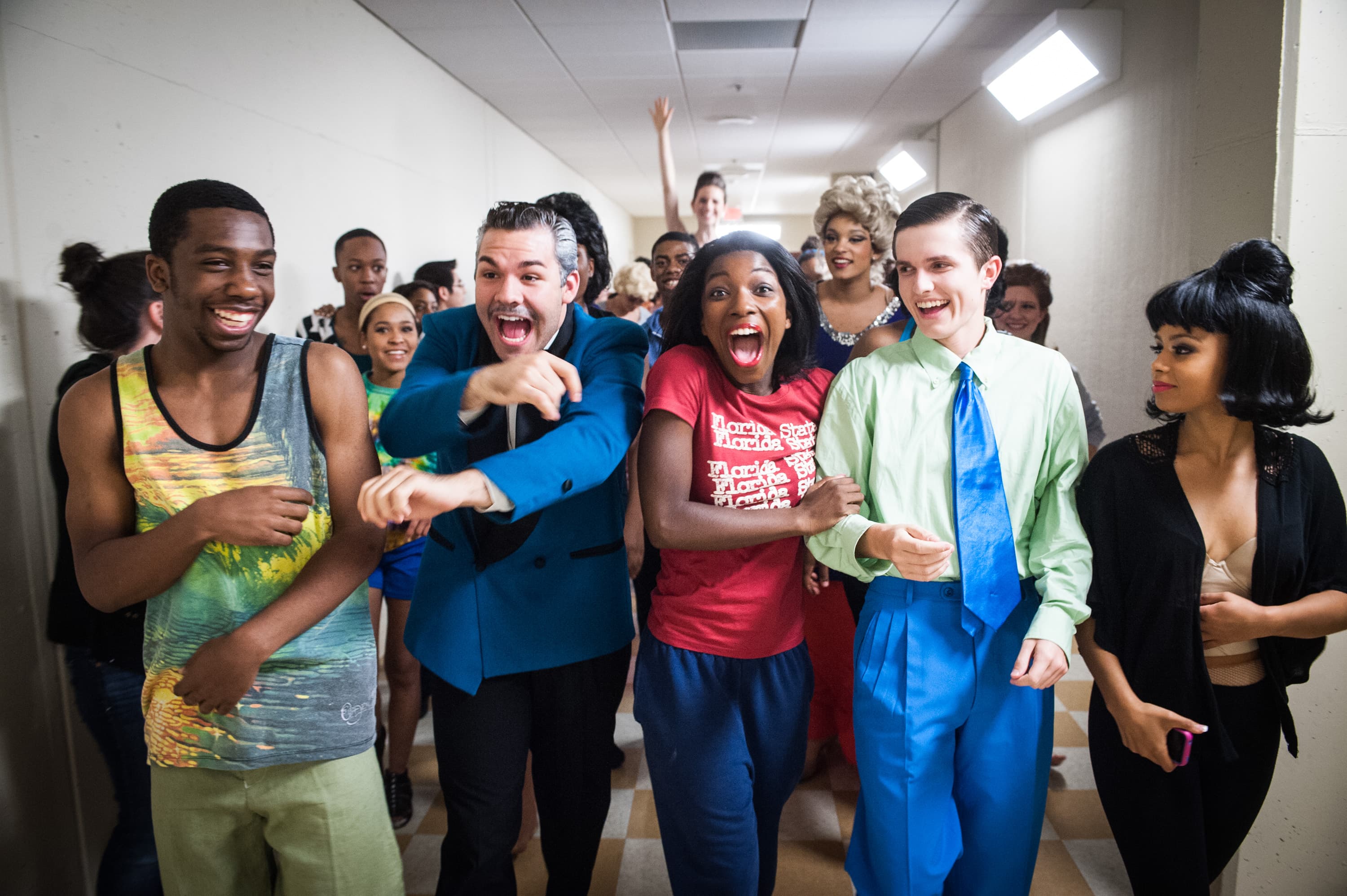 A group of people excitedly walking down a hallway. Some are wearing colorful outfits, and one person is in a blue jacket, while another wears a green shirt with a blue tie. A woman in a red shirt appears excited in the center of the group.