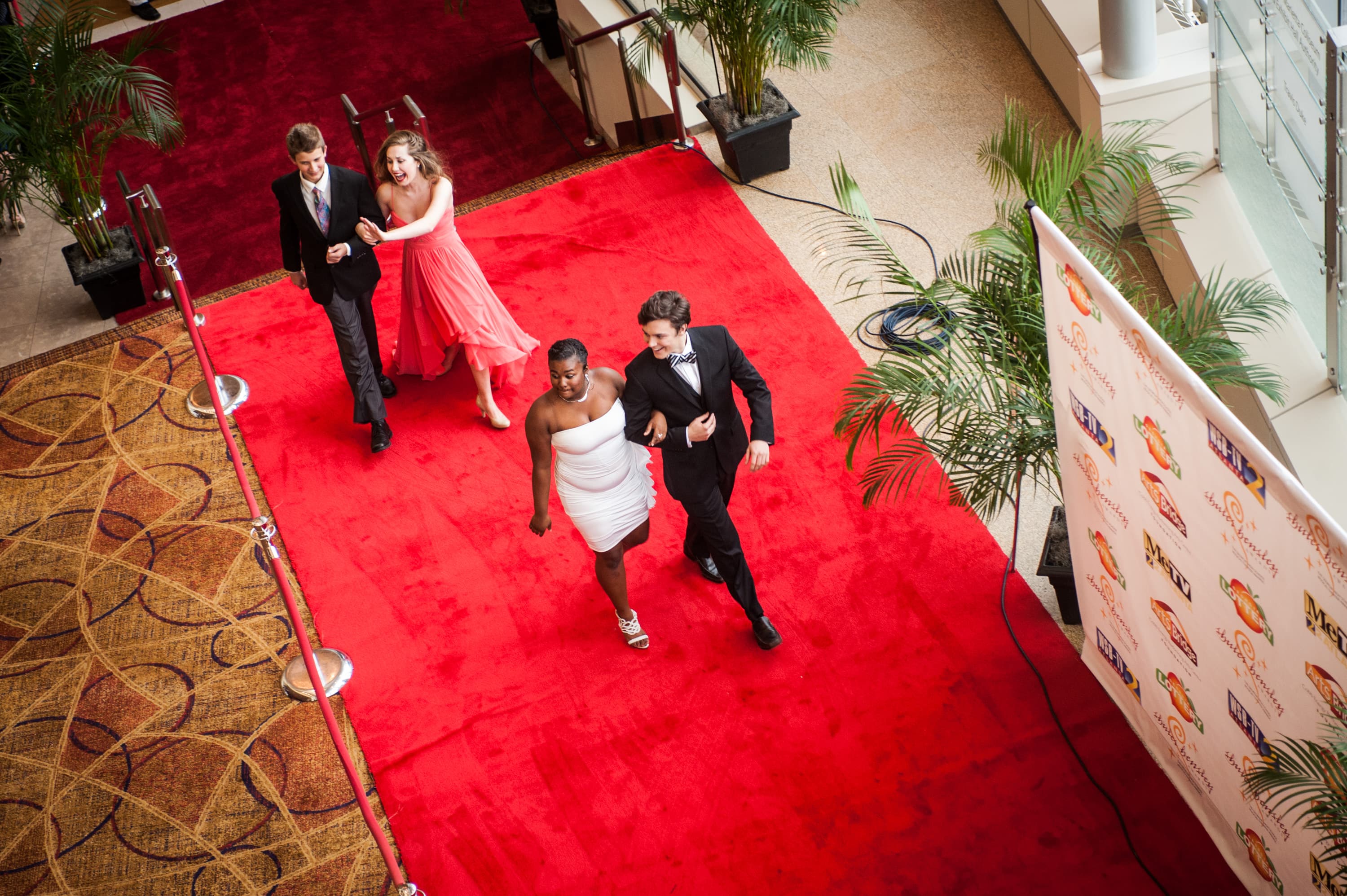 Four people walking on a red carpet, with two women in dresses and two men in suits, surrounded by red ropes and potted plants. A step and repeat banner is on the side displaying various logos.