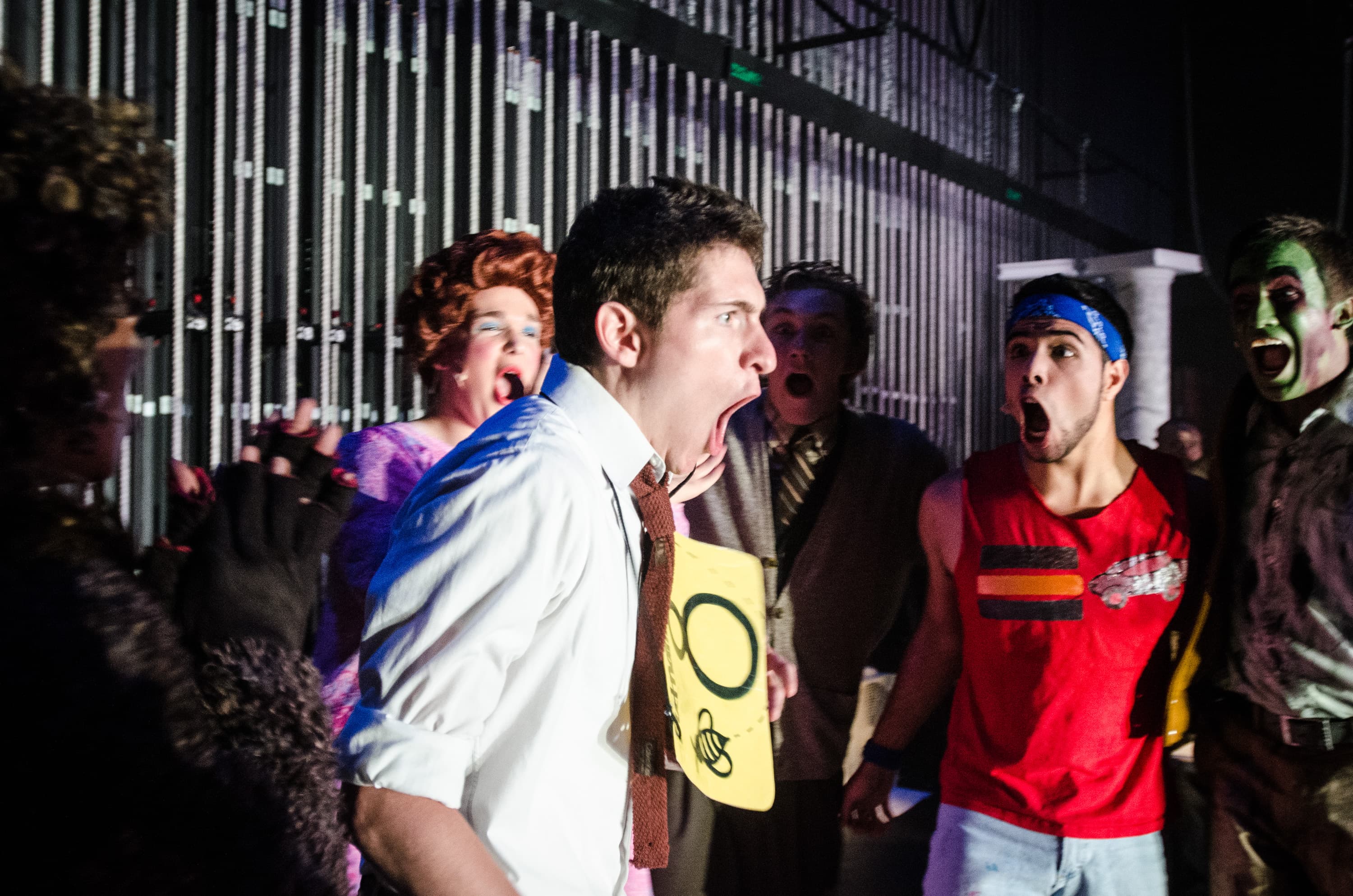 A group of people backstage in a theatrical setting, expressing surprise or excitement. One person in the front is wearing a white shirt and tie, holding a yellow sign with a bee drawing. Others in the background appear in varied costumes, including a person with a blue headband and another with face paint.